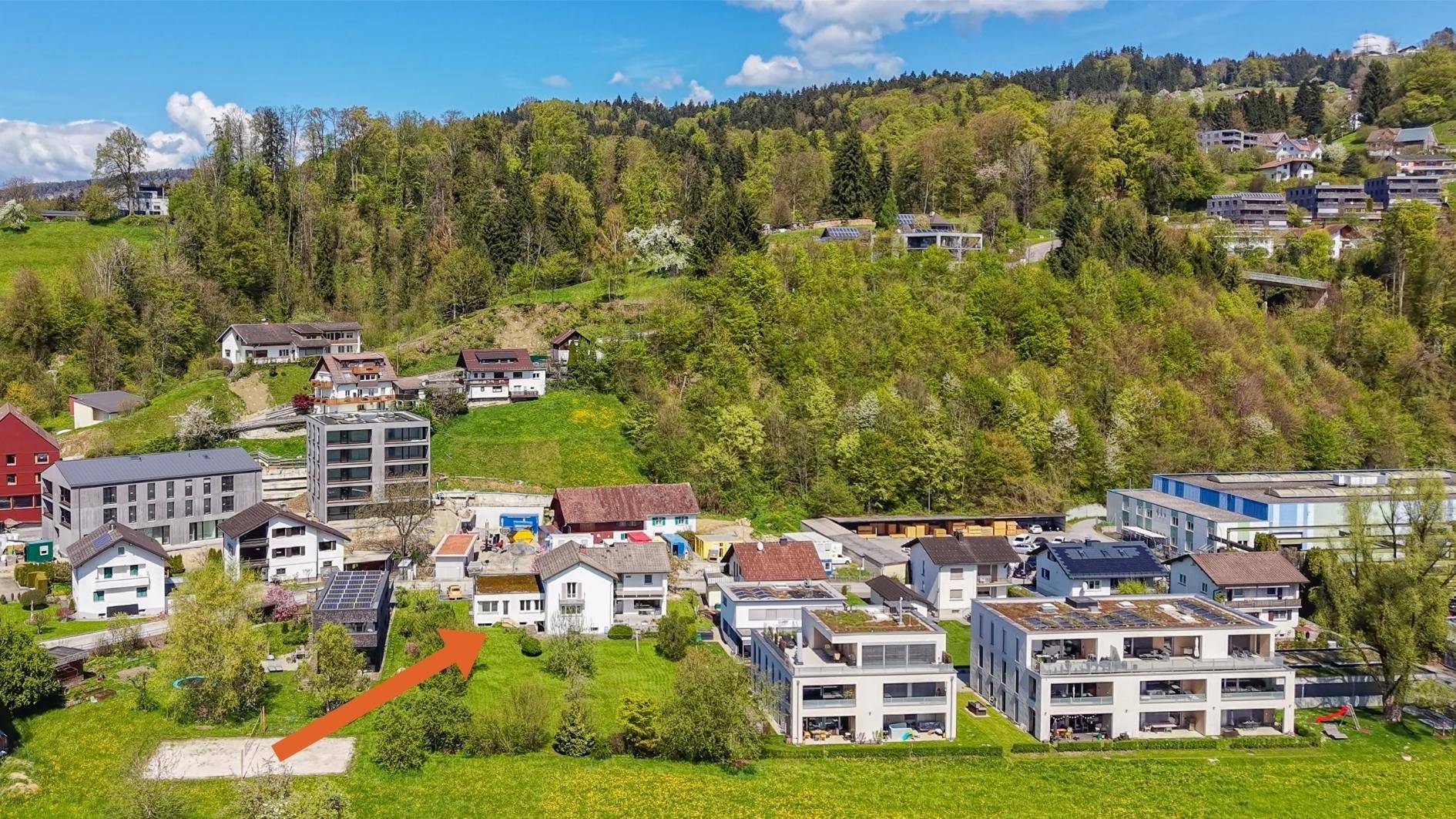 Luftaufnahme eines Wohngebiets mit Häusern und grünen Hügeln, ein Pfeil zeigt auf ein Grundstück.