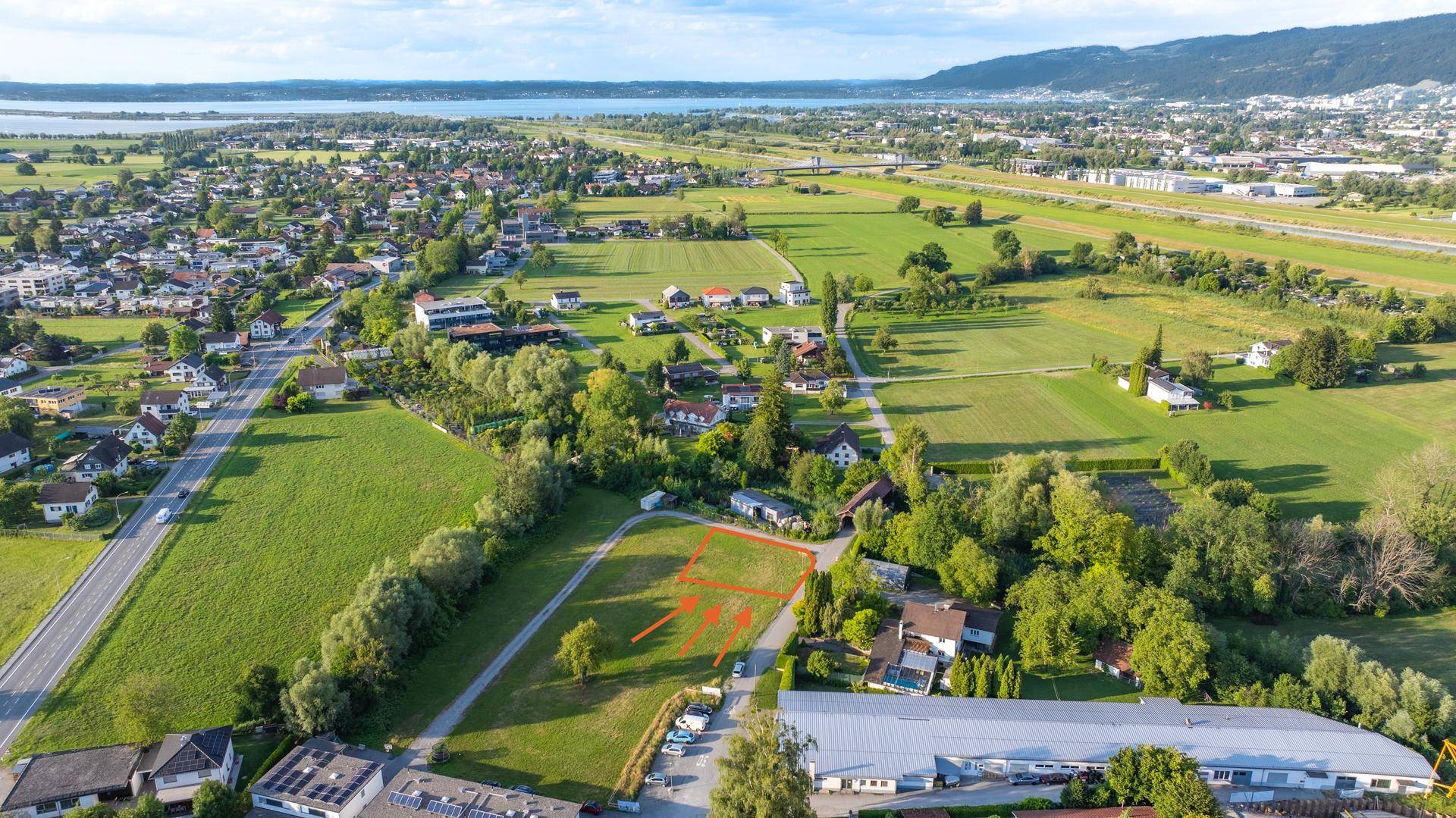 Luftaufnahme eines Baugrundstücks mit roter Markierung und Pfeilen, umgeben von Feldern und einem See in der Ferne.