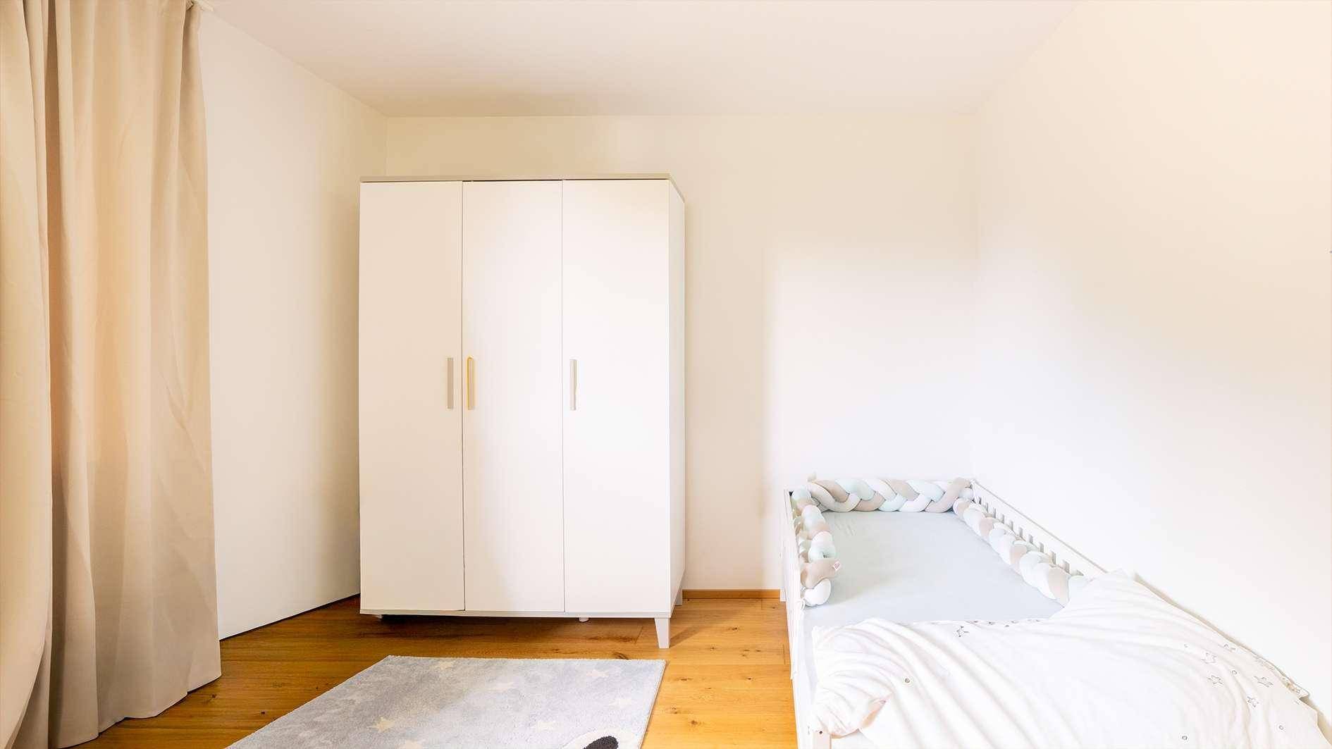Kinderzimmer mit Babybett, weißem Kleiderschrank und Vorhängen in neutralen Farben.
