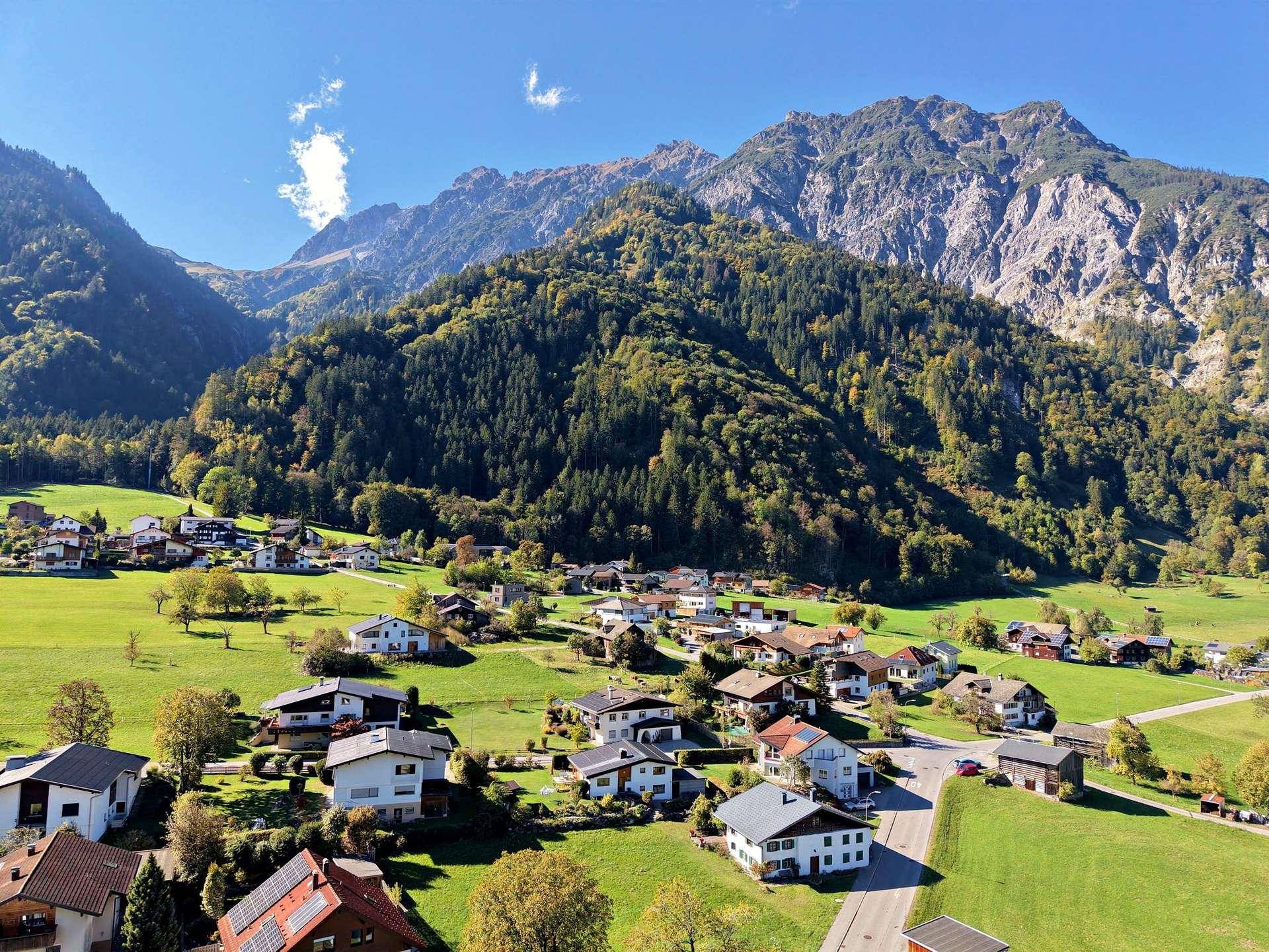 Luftaufnahme einer Siedlung mit vielen Häusern und bewaldeten Bergen im Hintergrund.