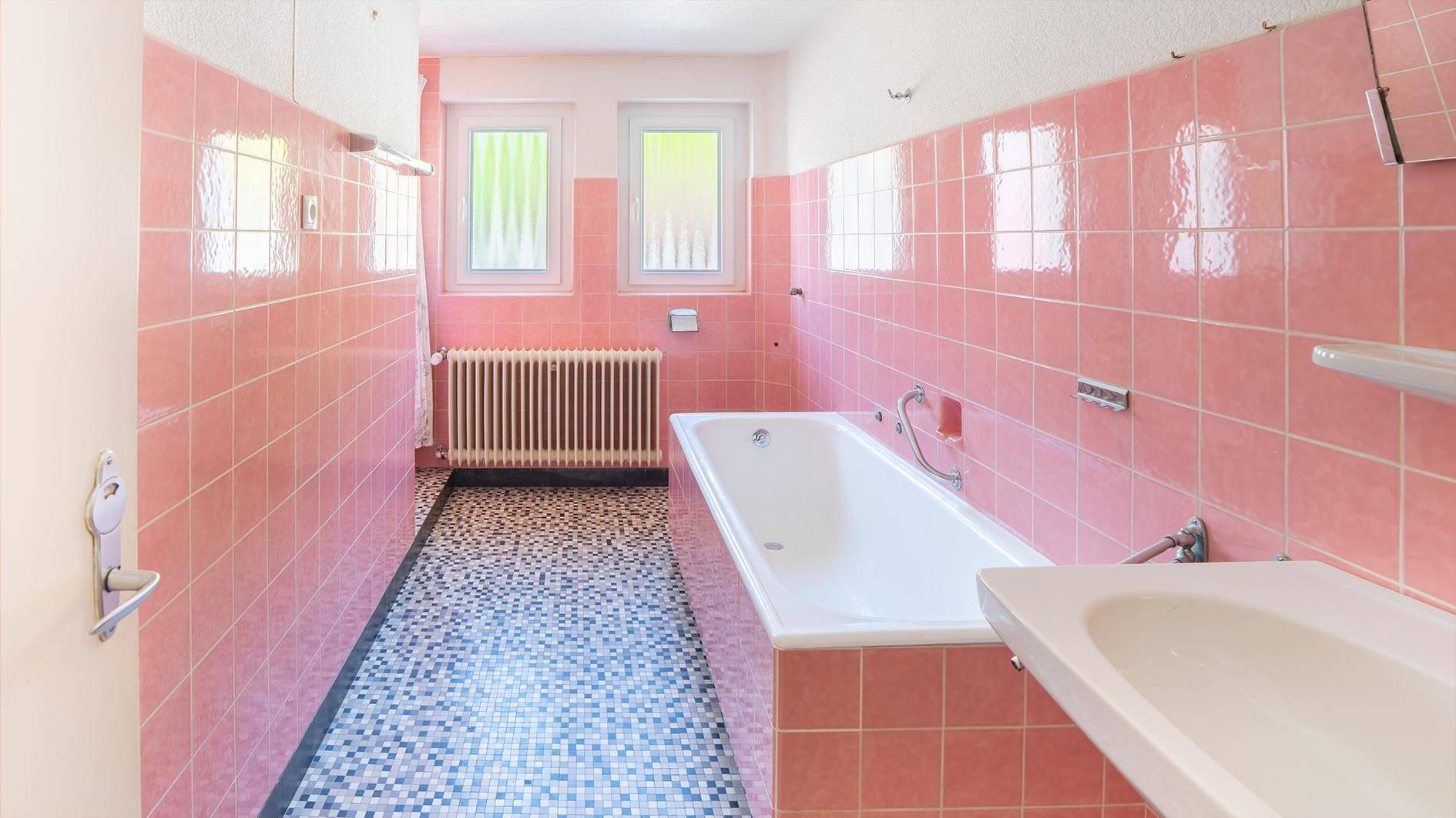Badezimmer im 80er-Jahre-Stil mit rosa Fliesen, Badewanne, Waschbecken und zwei Fenstern.