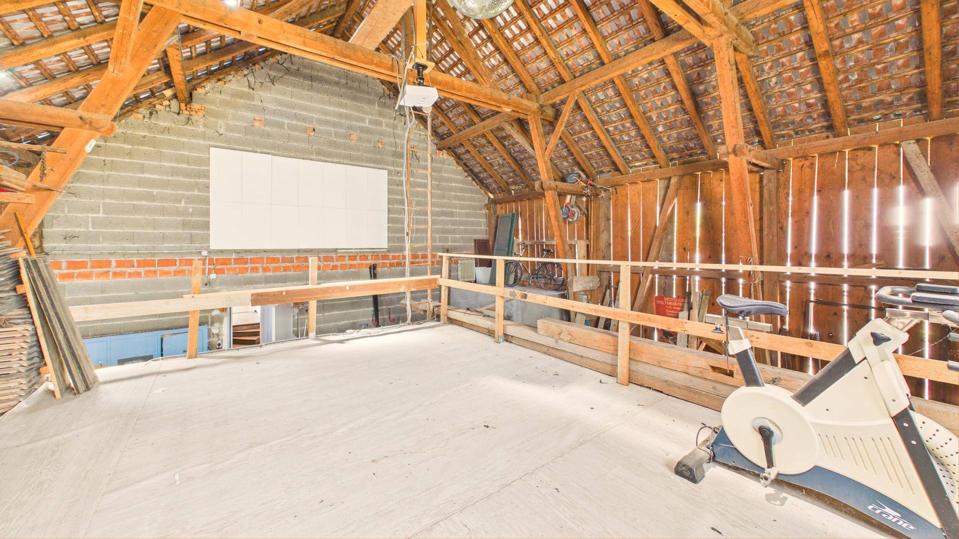 Ausbaufähiger Dachboden mit sichtbaren Holzbalken und Betonboden, als Fitnessbereich genutzt.