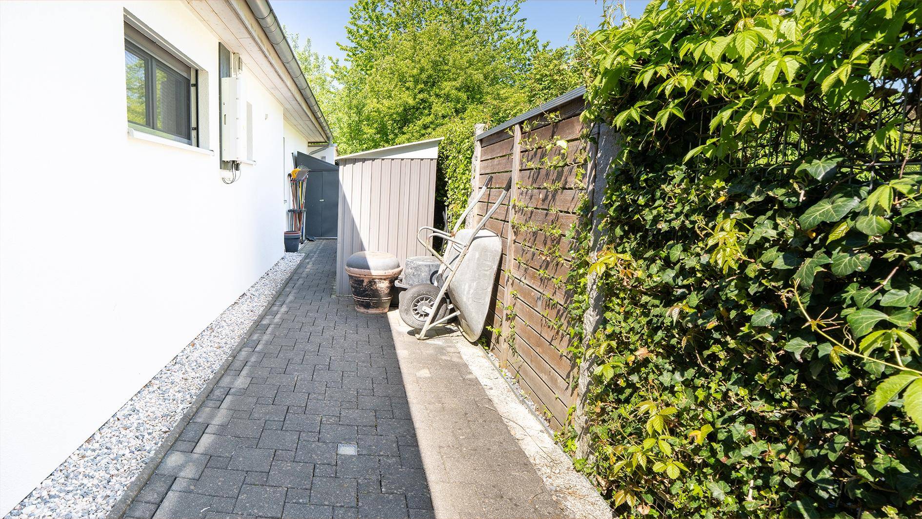 Schmaler gepflasterter Weg neben dem Haus, führt zu einem Geräteschuppen mit Efeu bewachsenem Zaun.