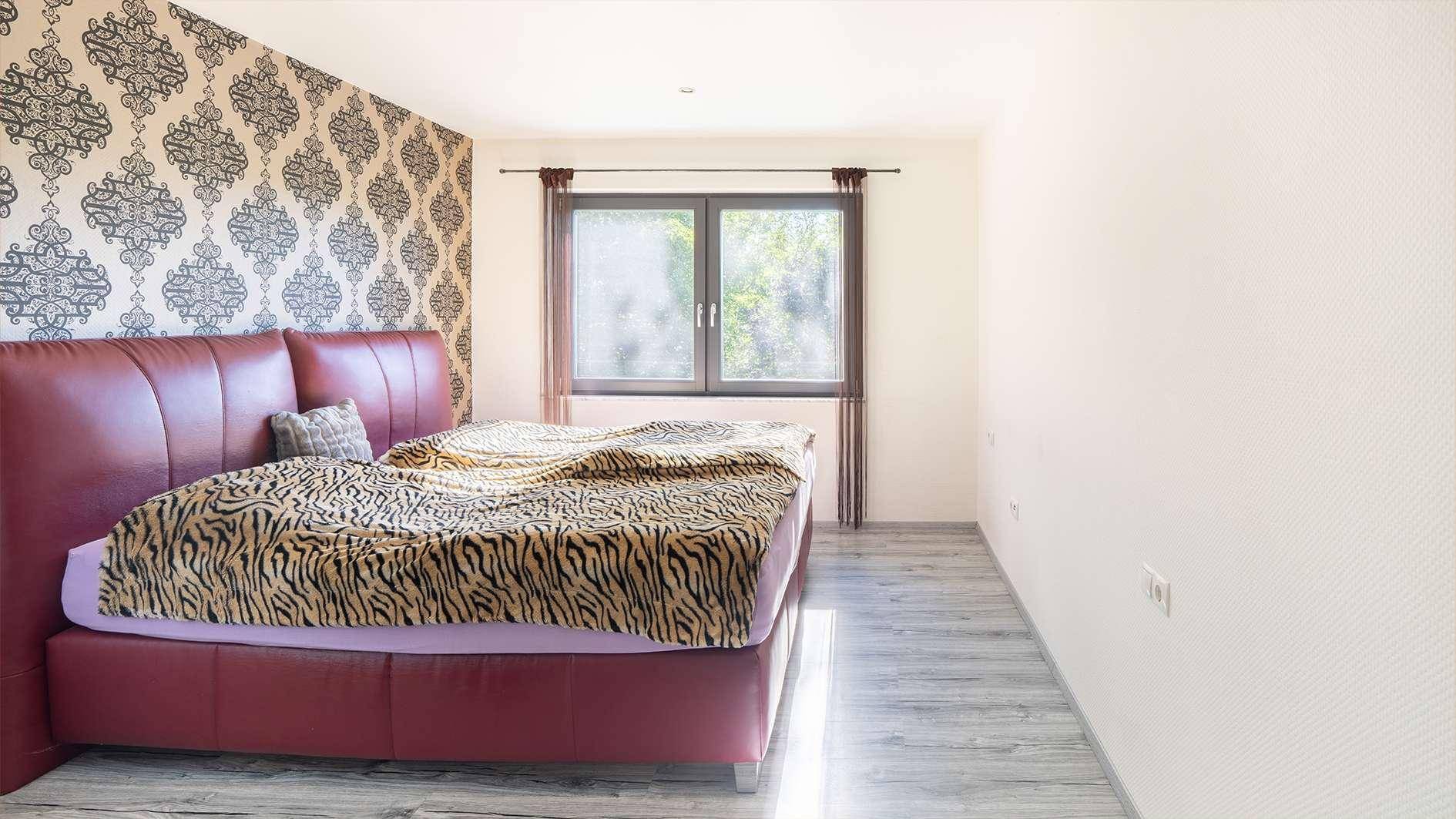 Helles Schlafzimmer mit rotem Doppelbett und gemusterter Akzentwand, bietet viel Tageslicht.