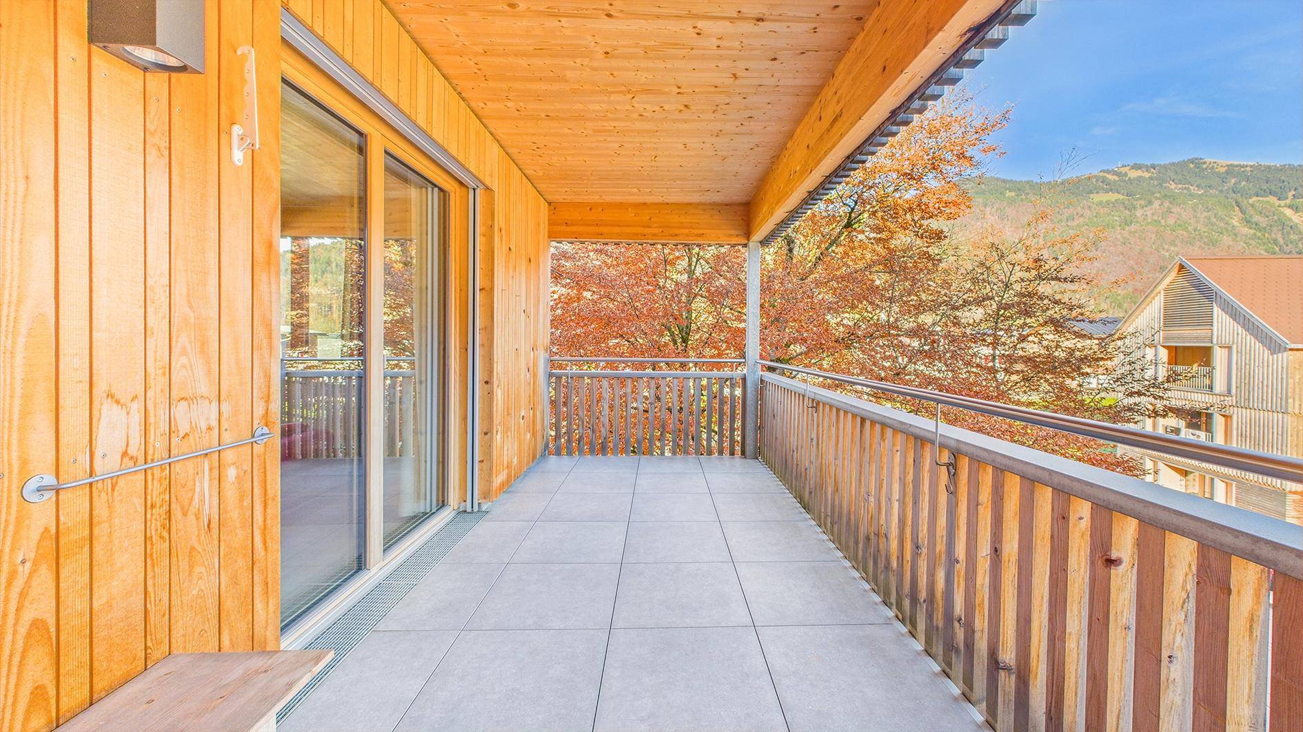 Langer Balkon mit Holzelementen, Fliesenboden und herbstlicher Aussicht auf die Umgebung.