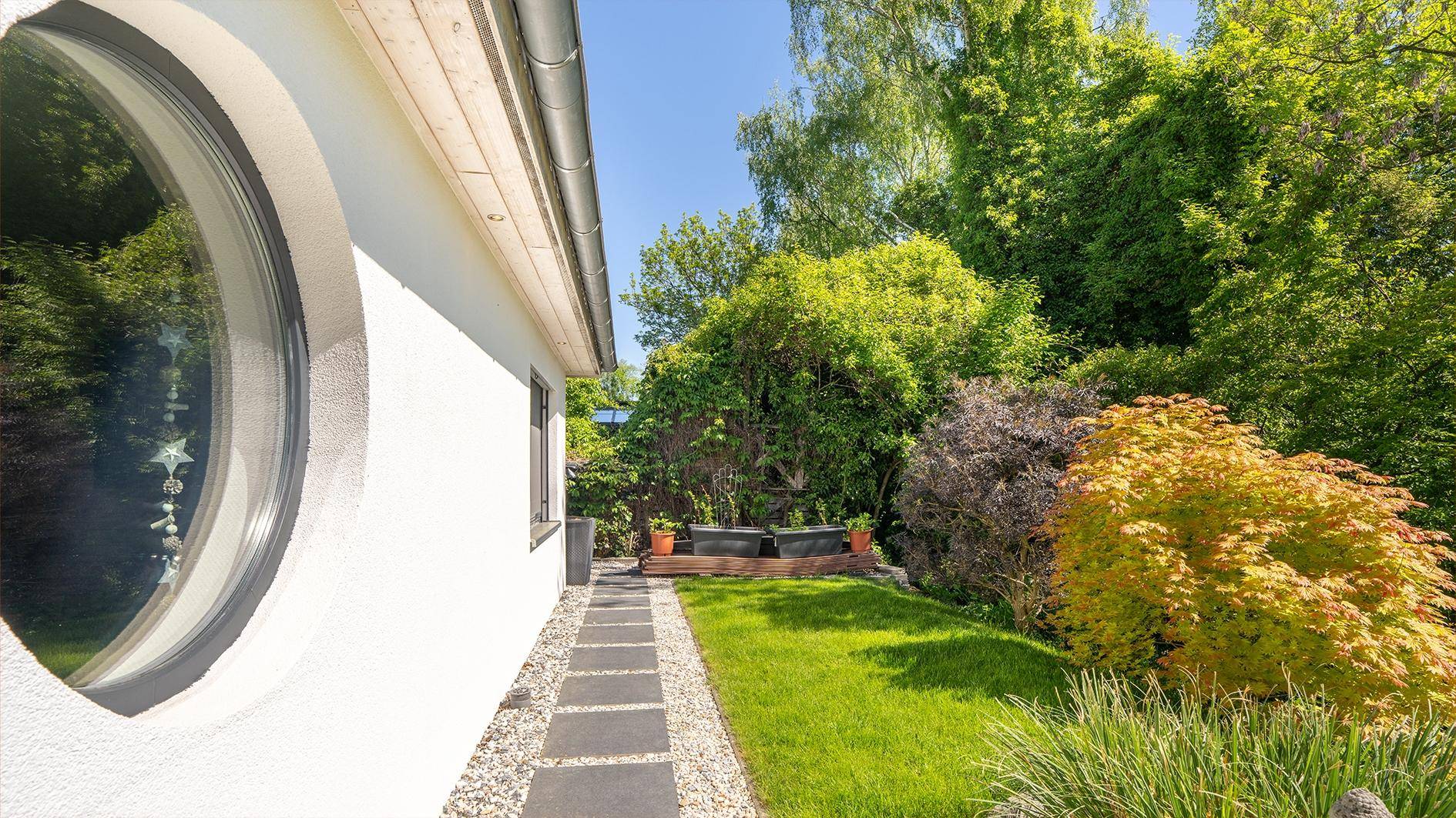 Seitlicher Gartenweg entlang des Hauses mit runden Fenstern und üppiger Bepflanzung.