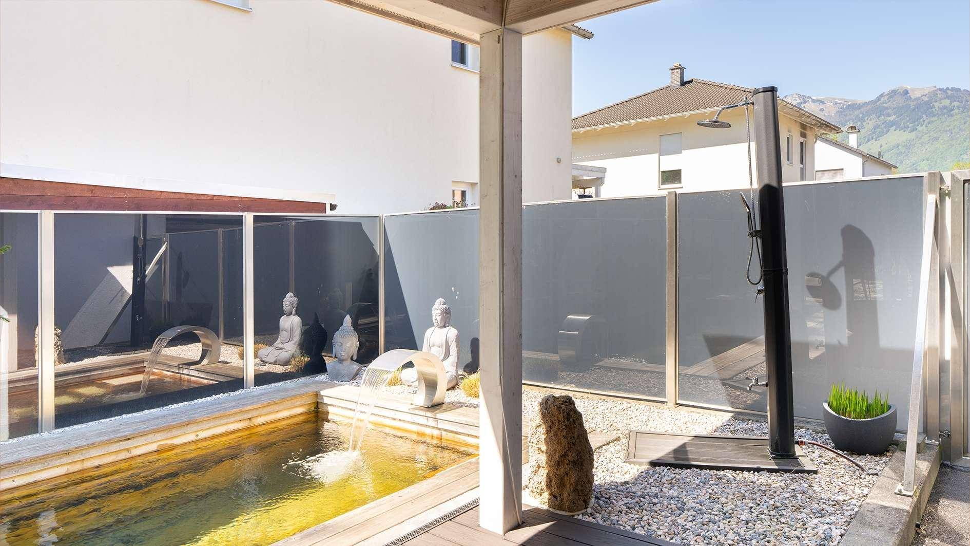 Außenbereich mit Dusche, dekorativem Teich und Buddha-Statuen, umgeben von Sichtschutz.