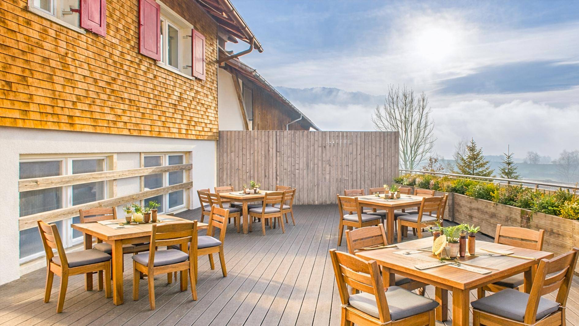 Große Holzterrasse mit mehreren Esstischen und Stühlen, bietet einen weiten Blick ins Grüne.
