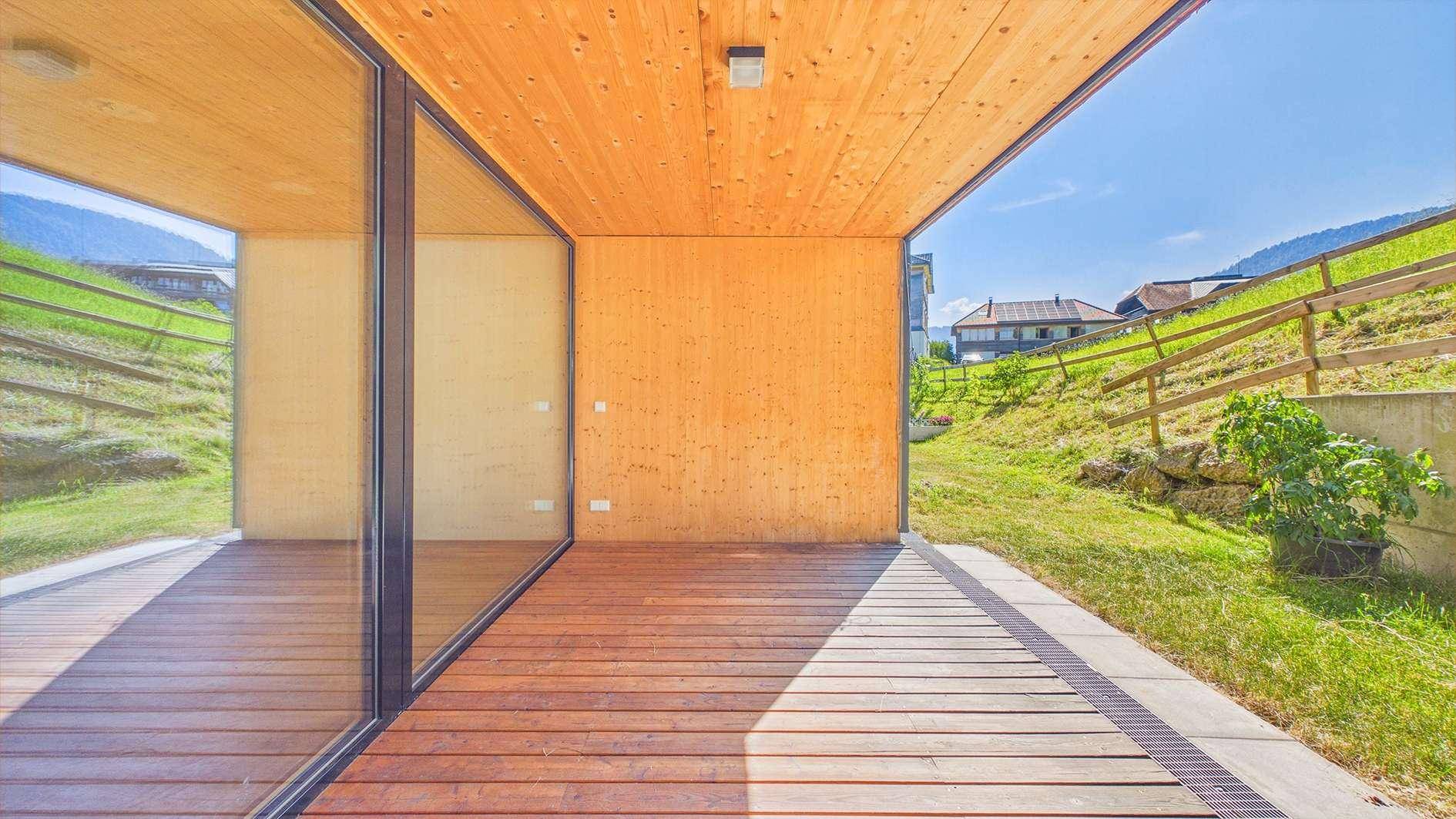 Sonniger Terrassenbereich mit Holzdeck und Holzdecke, umgeben von Glaswänden und Blick auf eine grüne Böschung.