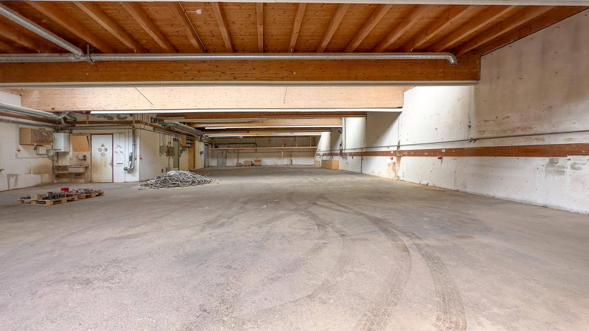 Großer, leerer Lagerraum mit Betonboden, sichtbaren Holzbalken und Kabeln, bereit zur Nutzung.