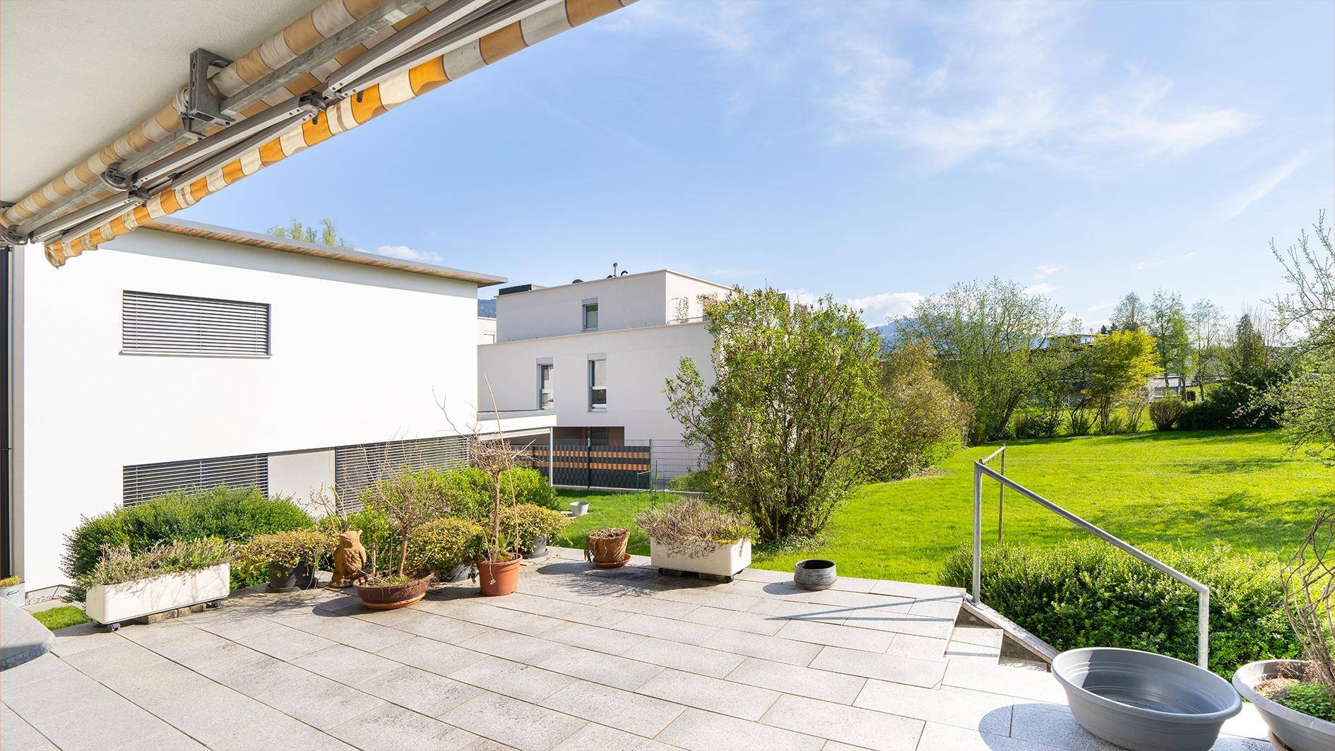 Große Terrasse mit grauen Fliesen, Topfpflanzen und Blick auf eine grüne Rasenfläche.
