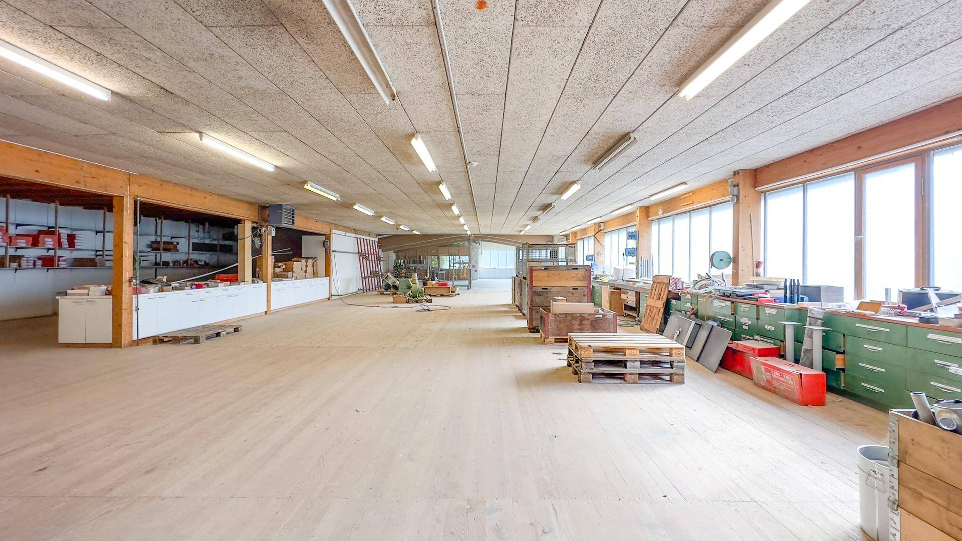 Geräumiger Werkstattbereich mit Holzfußboden, großen Fenstern und verschiedenen Arbeitsmaterialien und Geräten.