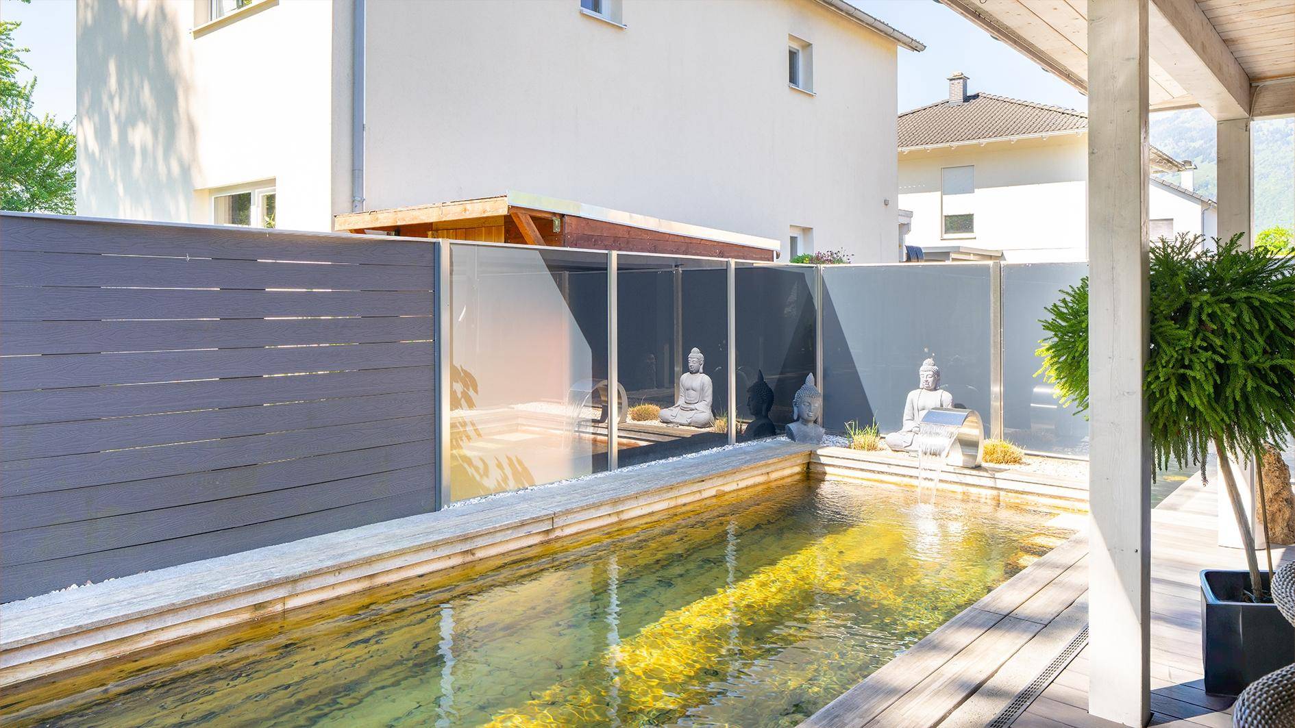 Gestalteter Gartenbereich mit Wasserbecken, Holzdeck und Sichtschutz, umgeben von grüner Natur.