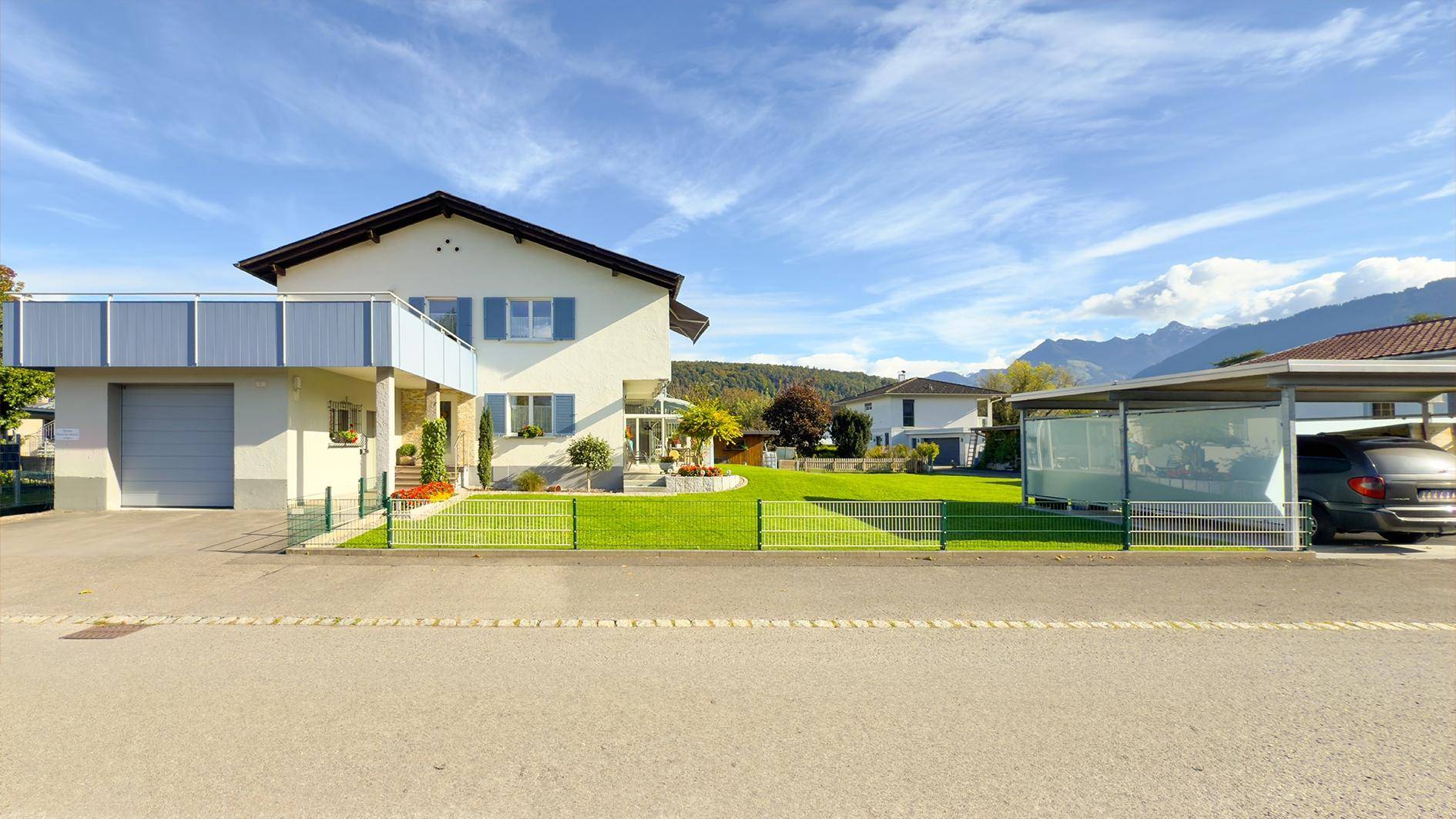 Freistehendes Einfamilienhaus mit großem Garten, Carport und Bergblick an einem sonnigen Tag.