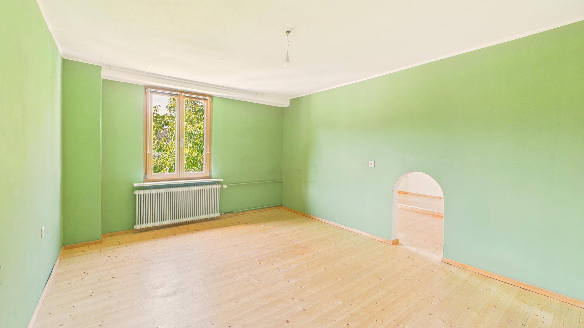 Helles Zimmer mit grünen Wänden, Holzboden und Fenster mit Blick ins Grüne.
