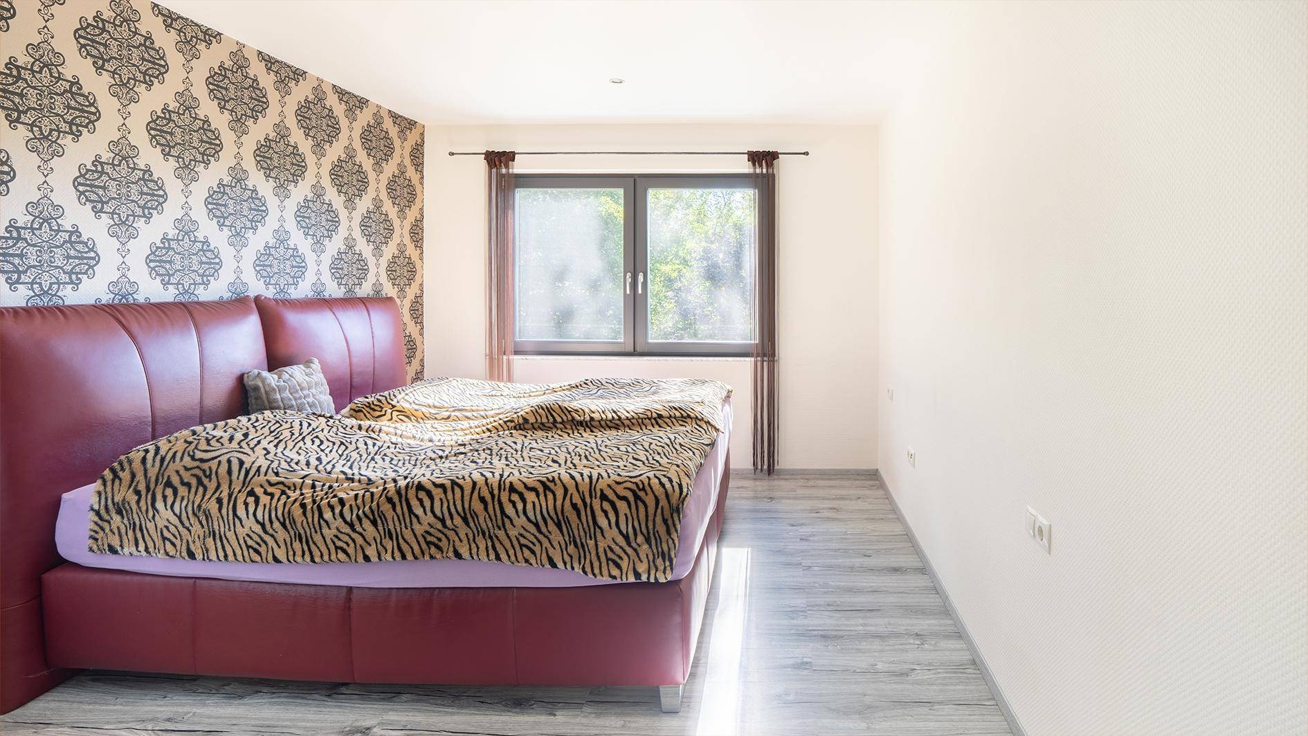 Schlafzimmer mit rotem Bett, gemusterter Wand und Fenster, das viel natürliches Licht hereinlässt.