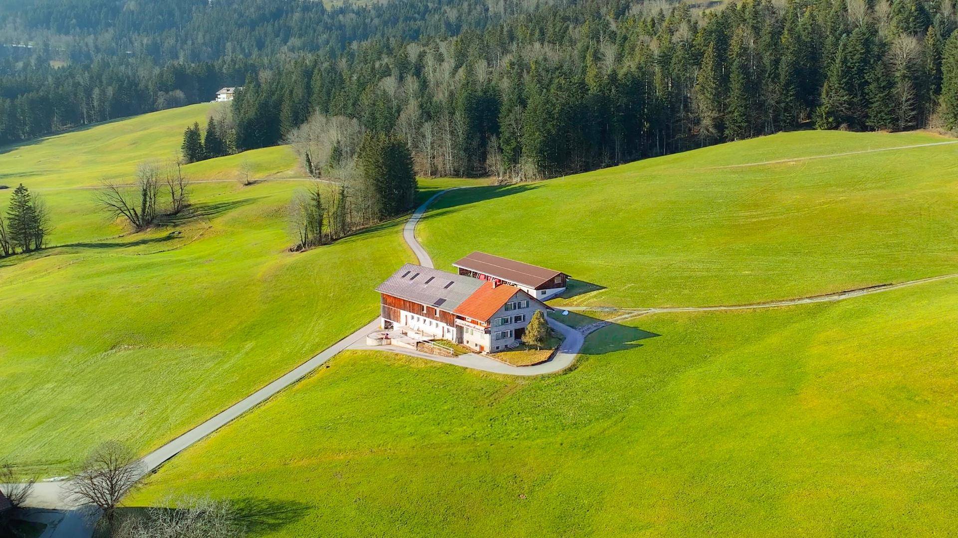 Weitläufige Luftaufnahme des Anwesens mit Bauernhaus und Nebengebäude inmitten grüner Hügel und Wälder.