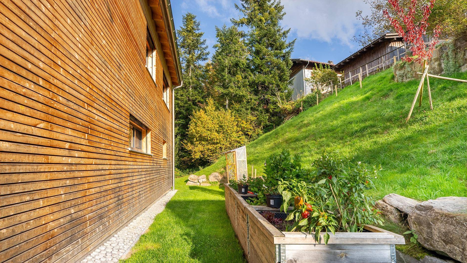 Seitenansicht des Hauses mit Holzfassade und einem Hochbeet im sonnigen Gartenbereich.