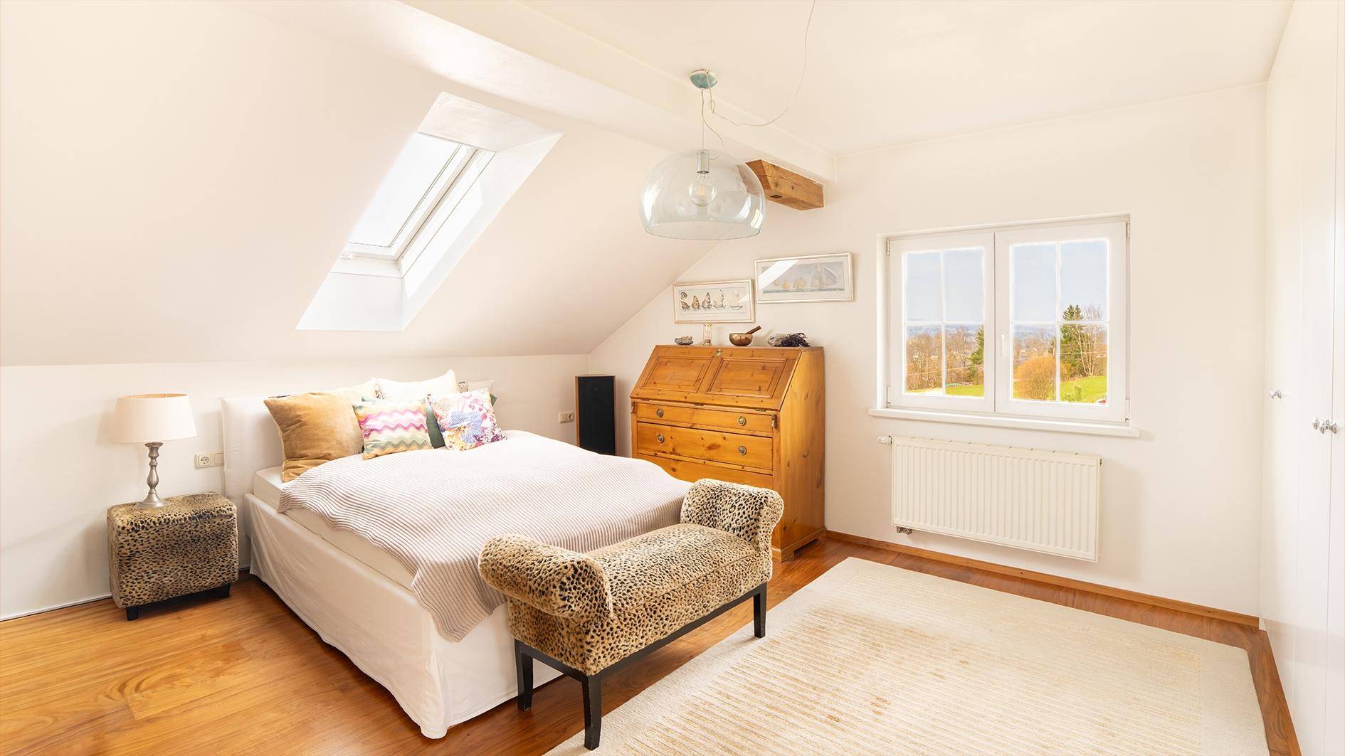 Helles Schlafzimmer im Dachgeschoss mit Holzboden, Dachfenster und Blick auf die grüne Umgebung.