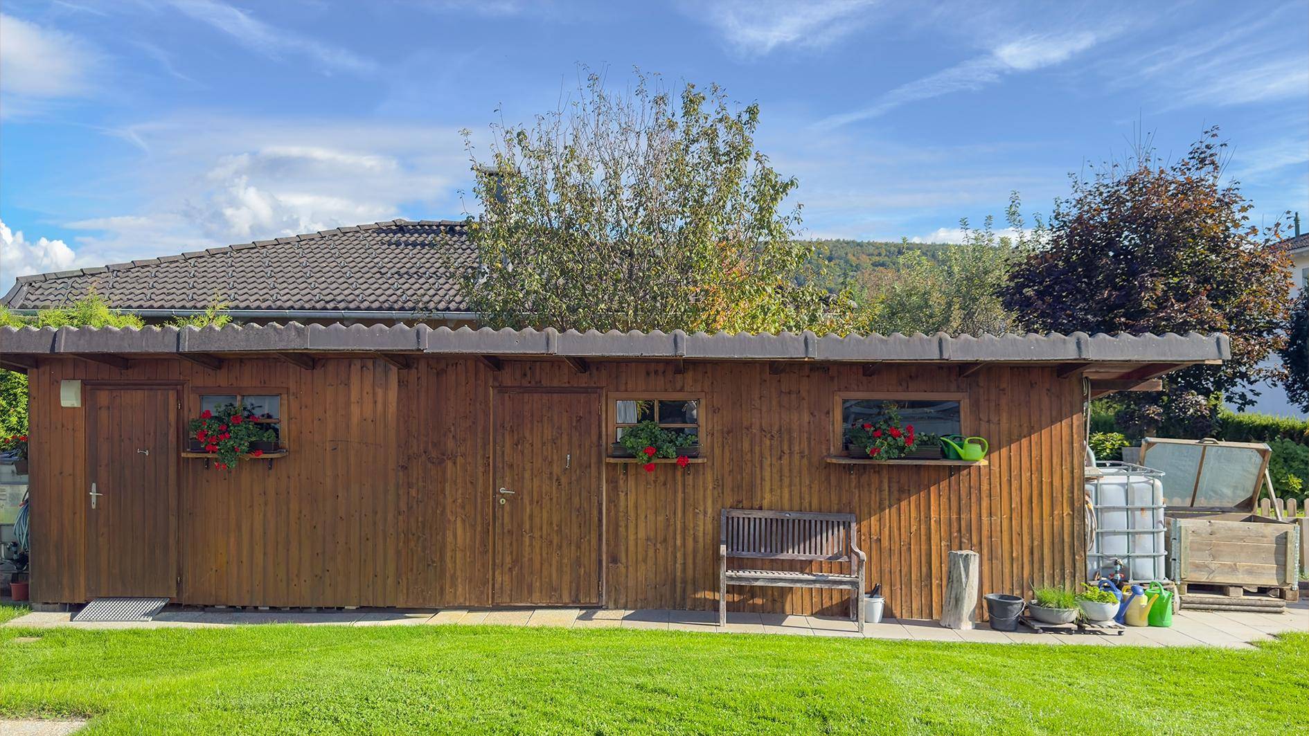 Rustikaler Holzschuppen im Garten mit Blumenkästen und Gartenbank unter blauem Himmel.