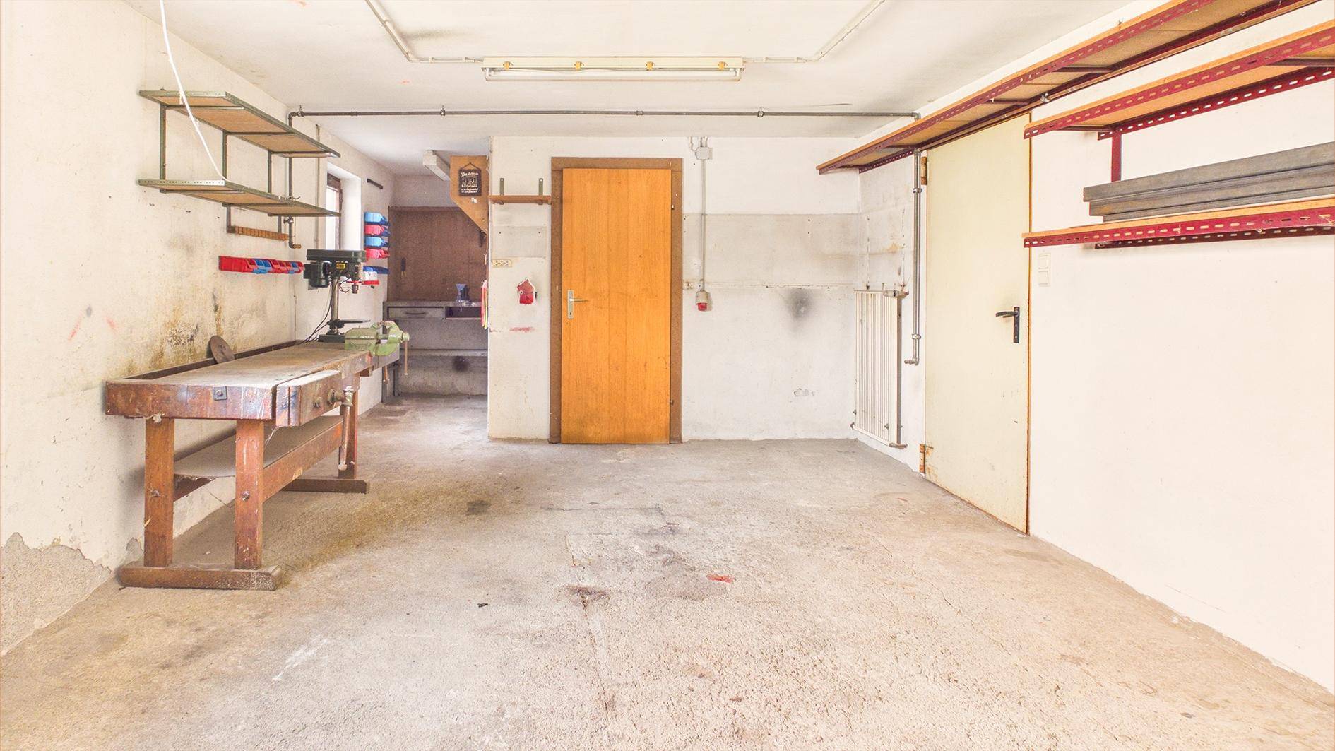 Geräumige Garage oder Werkstatt mit Betonboden, Werkbank und Regalen für Lagerung.