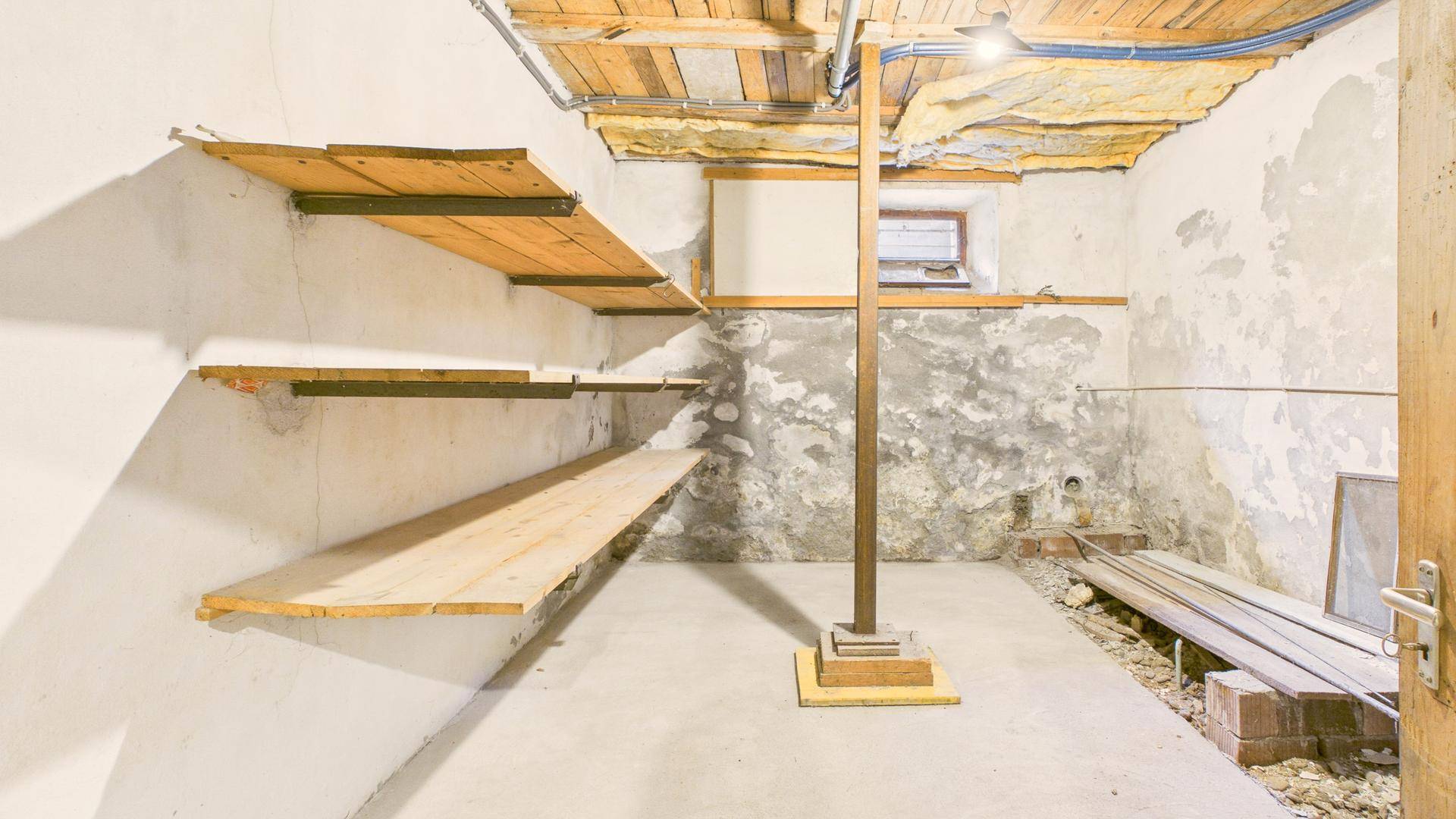 Unausgebauter Kellerraum mit Betonboden, Holzregalen und sichtbaren Leitungen.