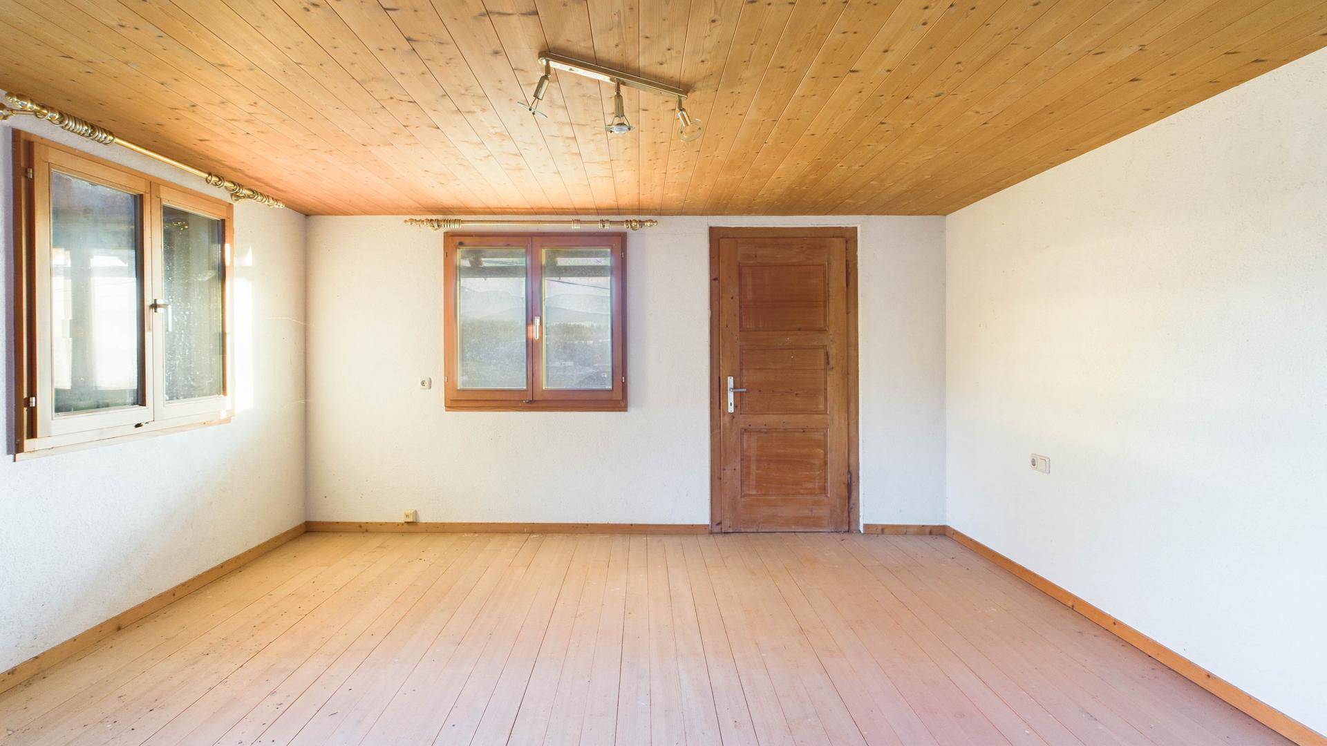 Leerer Raum mit Holzdielenboden, Holzdecke und zwei Fenstern, bereit zur Renovierung.