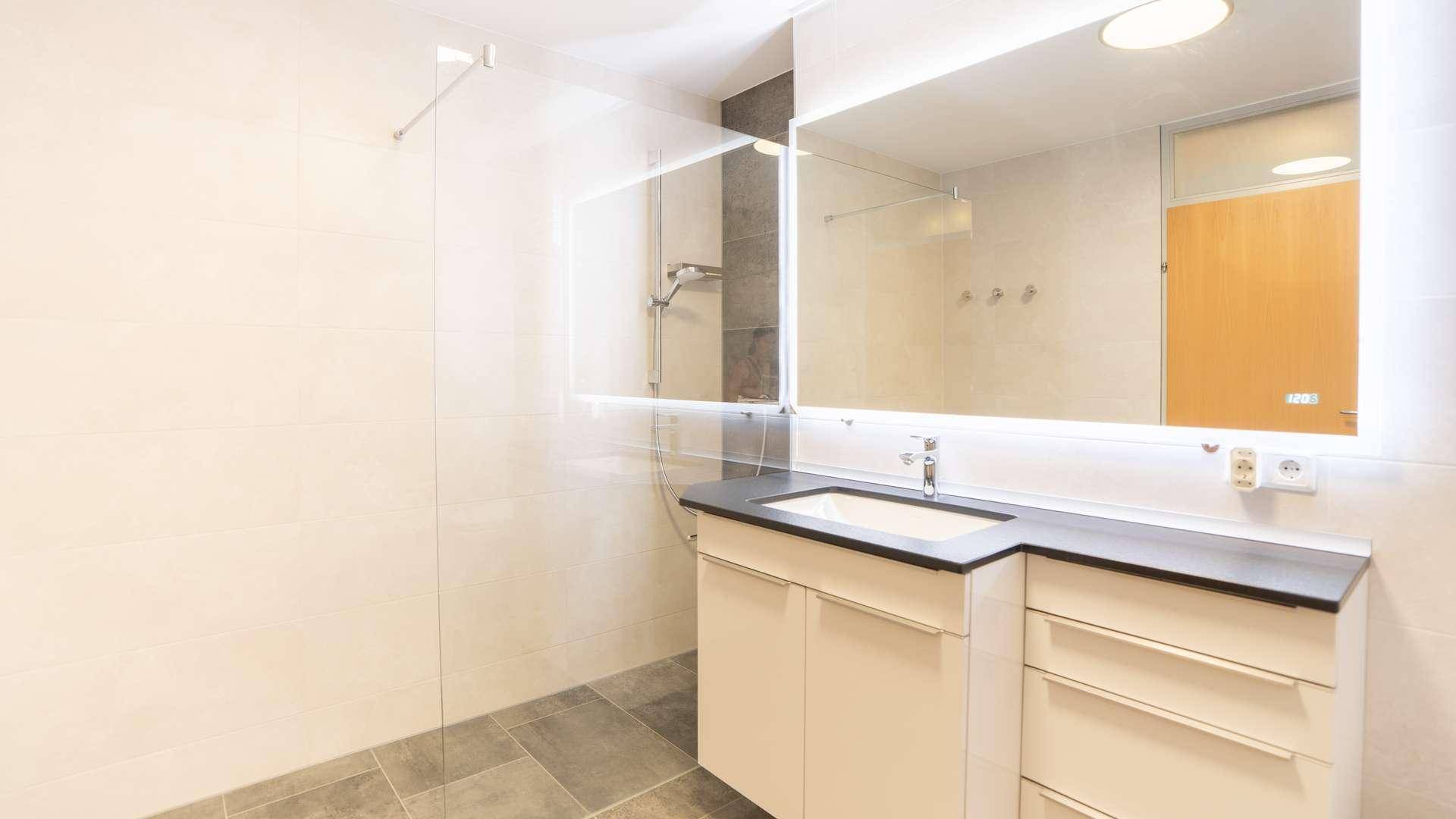 Modernes Badezimmer mit begehbarer Dusche, großem Spiegel und eleganten Fliesen in Hell- und Dunkelgrau.
