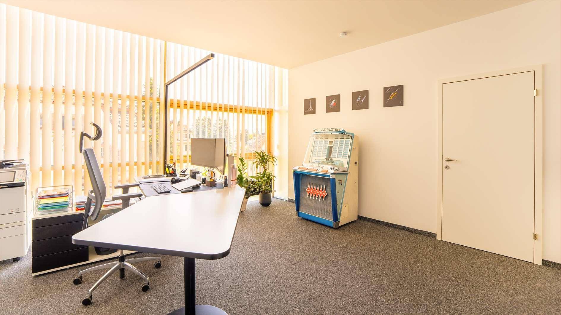 Modernes Büro mit Schreibtisch, Computer und einer einzigartigen Jukebox, beleuchtet durch große Fenster.