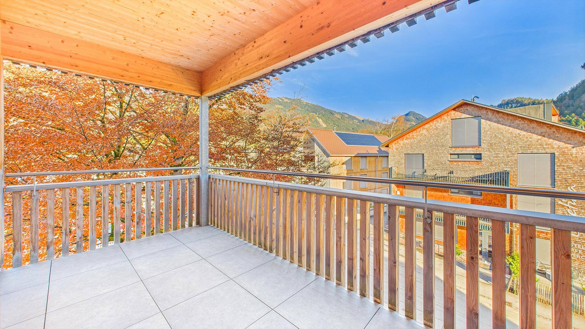 Geräumiger Balkon mit Holzelementen, Fliesenboden und Blick auf die umliegenden Gebäude und Berge.