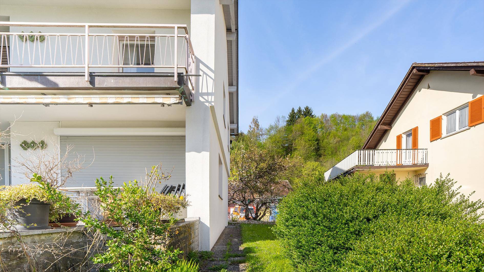 Seitenansicht eines weißen Gebäudes mit Balkon und grüner Hecke, angrenzend an ein Nachbarhaus.