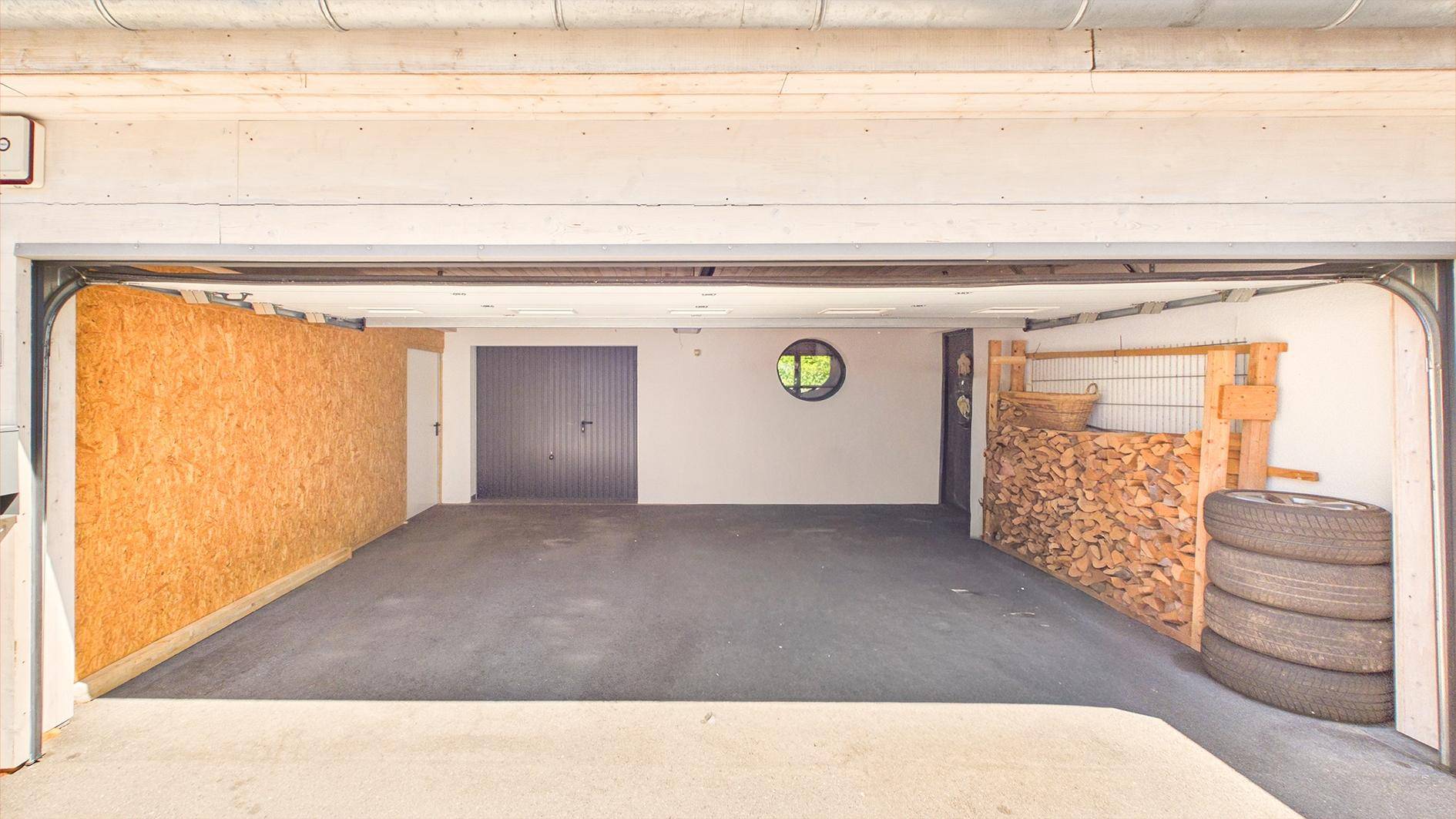 Geräumige Garage mit Betonboden, rundem Fenster und Lagerplatz für Holz und Reifen.