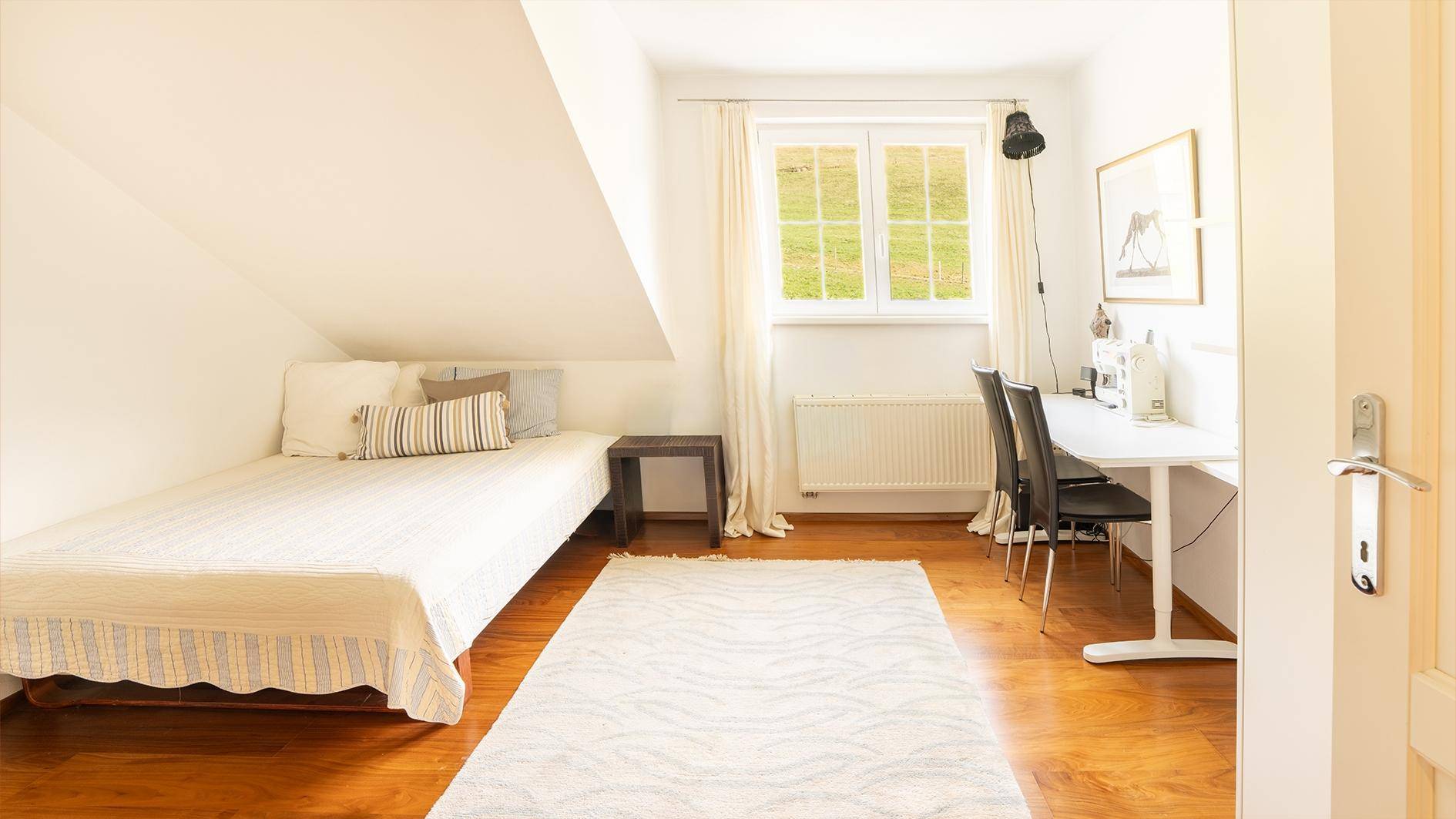 Helles Gästezimmer mit Einzelbett, Schreibtisch und Fenster mit Blick auf die grüne Landschaft.