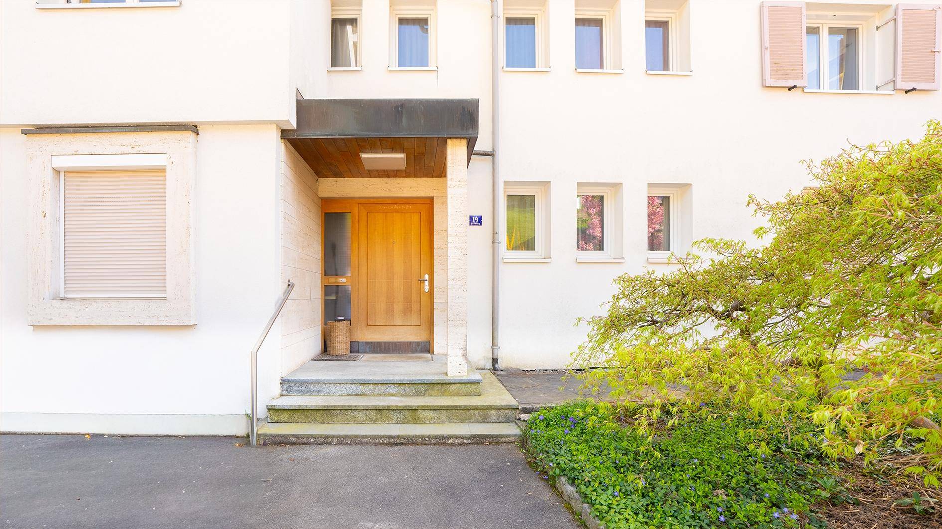 Eingangsbereich eines weißen Gebäudes mit Holzhaustür, Steinstufen und grüner Bepflanzung.