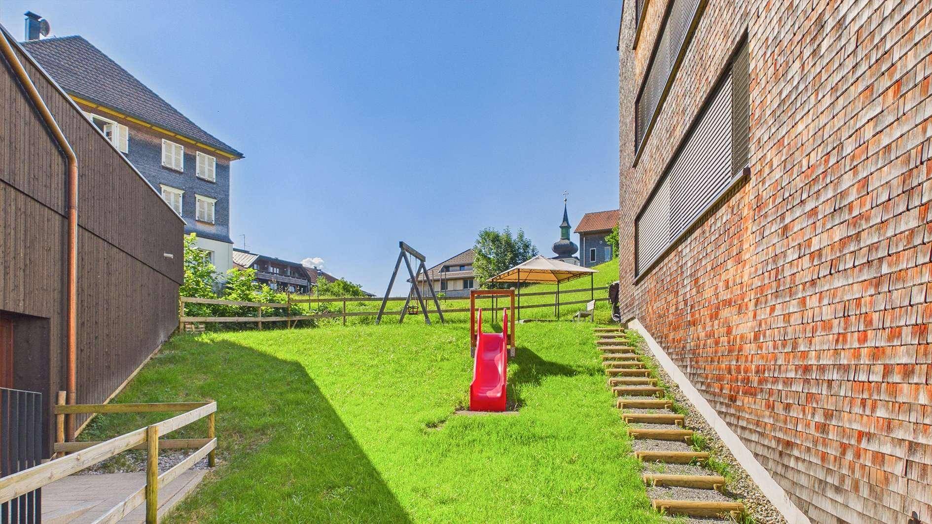 Grüner Gartenbereich mit Kinderspielplatz, Rutsche und Schaukel, neben dem Gebäude mit Holzschindelfassade.