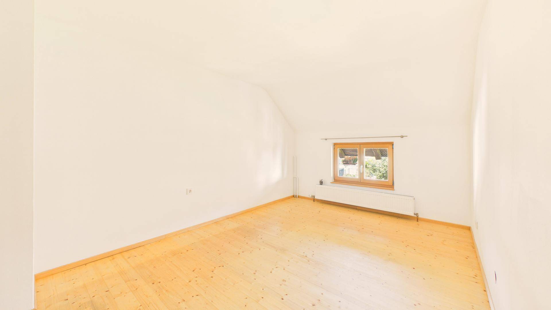Helles Zimmer mit Holzboden, Dachschräge und Fenster mit Blick ins Grüne.