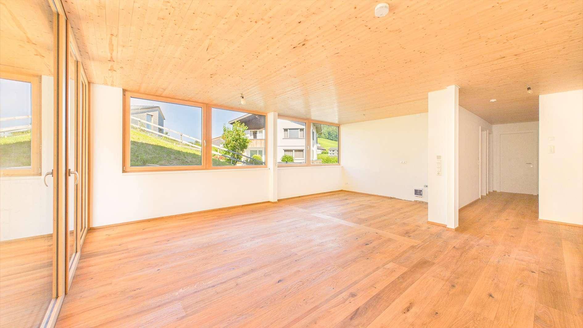 Leerer, heller Raum mit Holzboden, großen Fenstern und Zugang zu einem Außenbereich.