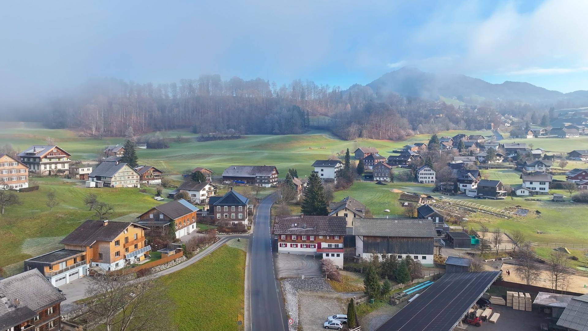 Weitläufige Luftaufnahme eines malerischen Dorfes in einer grünen Hügellandschaft mit Bergen im Hintergrund.