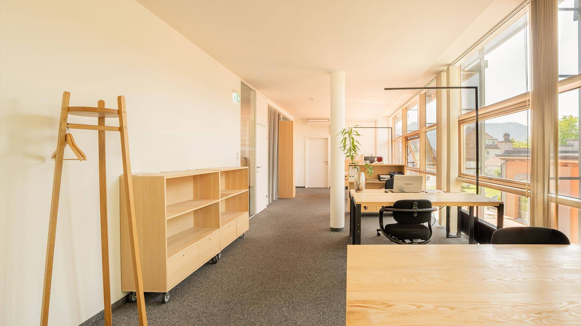 Heller Bürobereich mit großen Fenstern, Teppichboden und modernen Holzmöbeln, bietet viel Tageslicht.