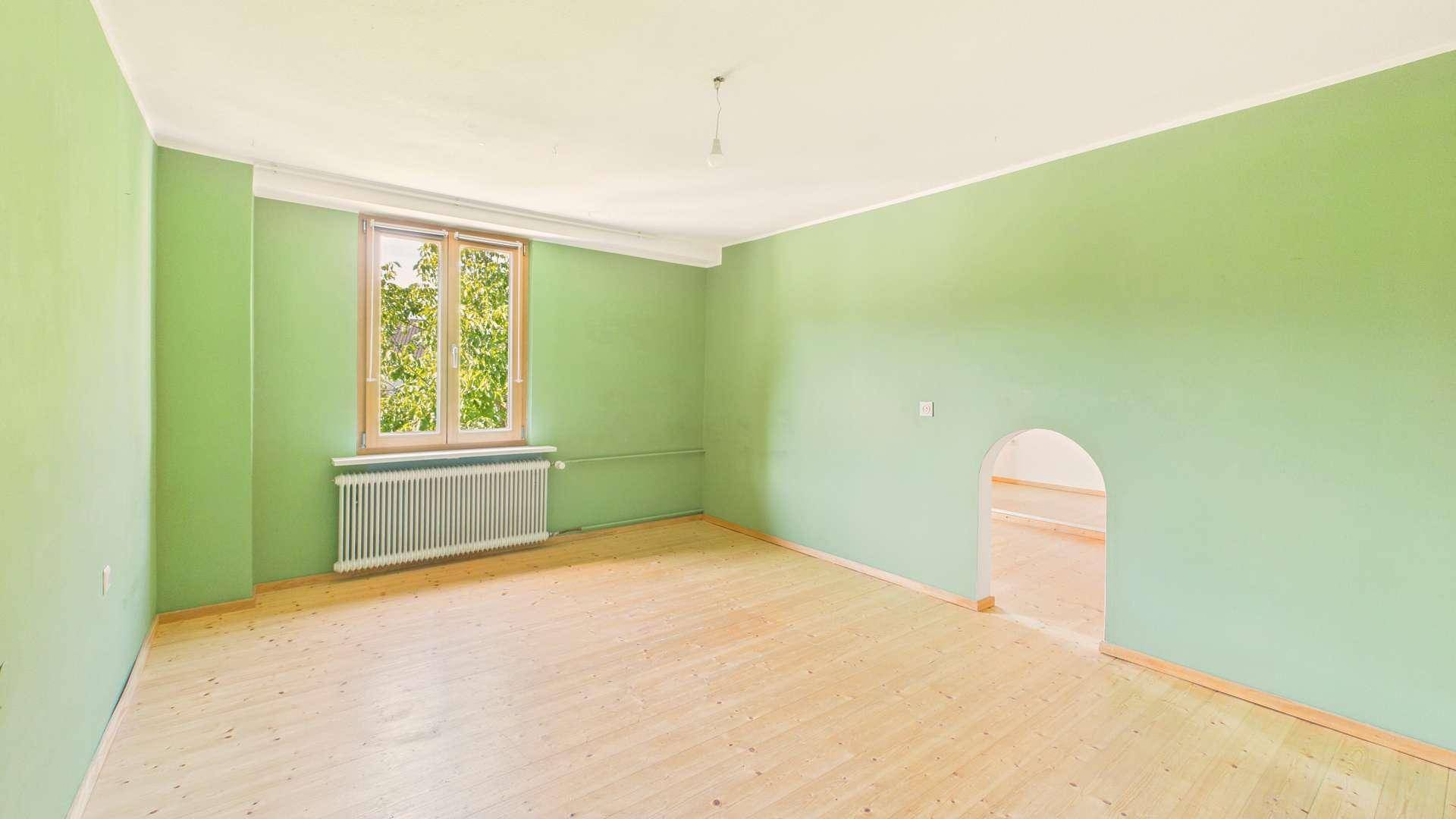 Geräumiges Zimmer mit grünen Wänden, Holzboden und Fenster zum Garten.
