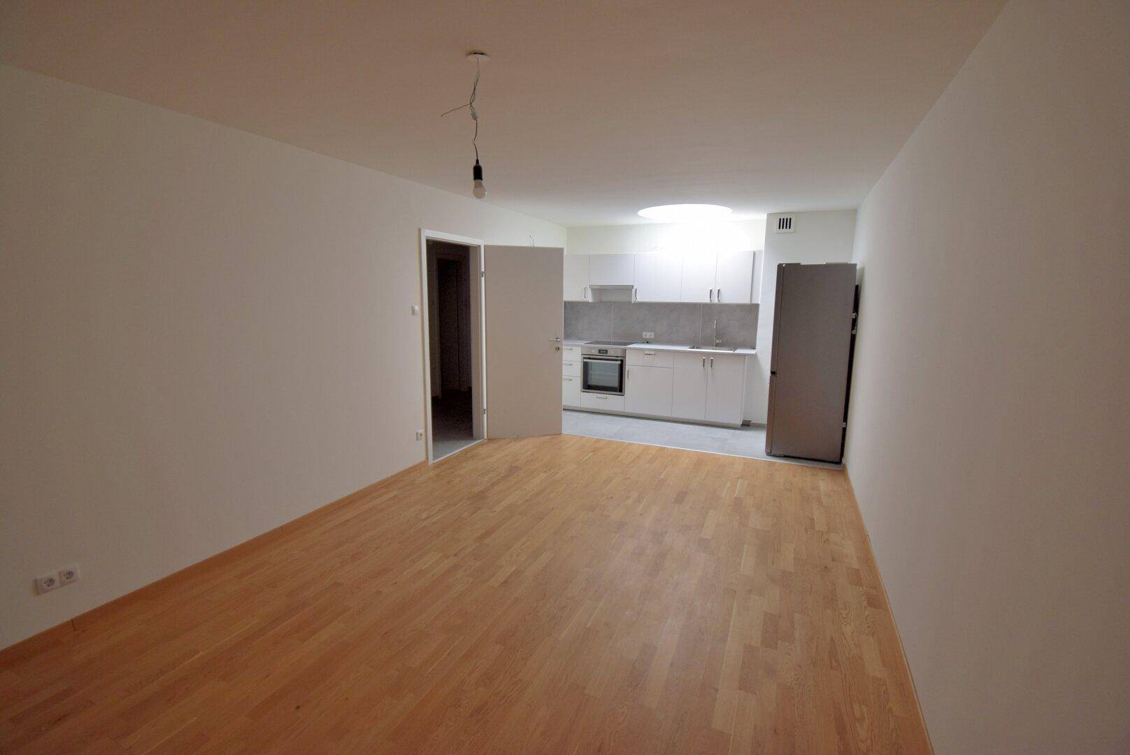 Geräumiges Wohnzimmer mit hellem Parkettboden und Blick auf die offene Küche.