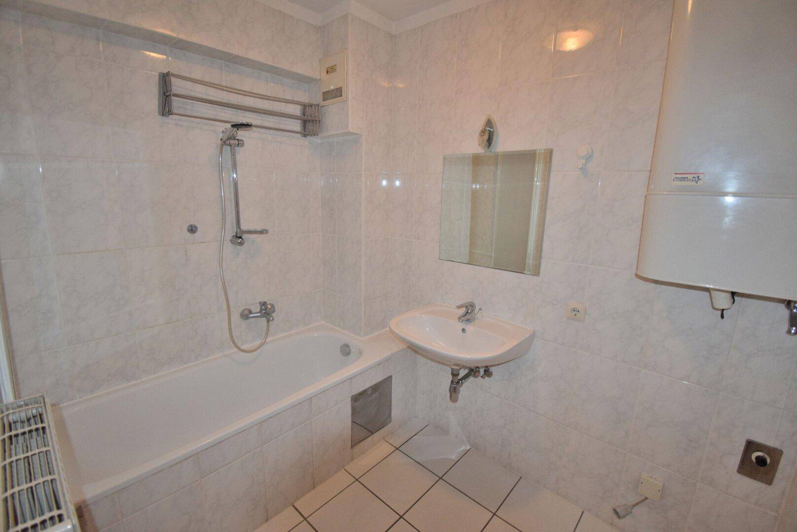Badezimmer mit weißen Fliesen, einer Badewanne, Waschbecken und einem Warmwasserboiler.