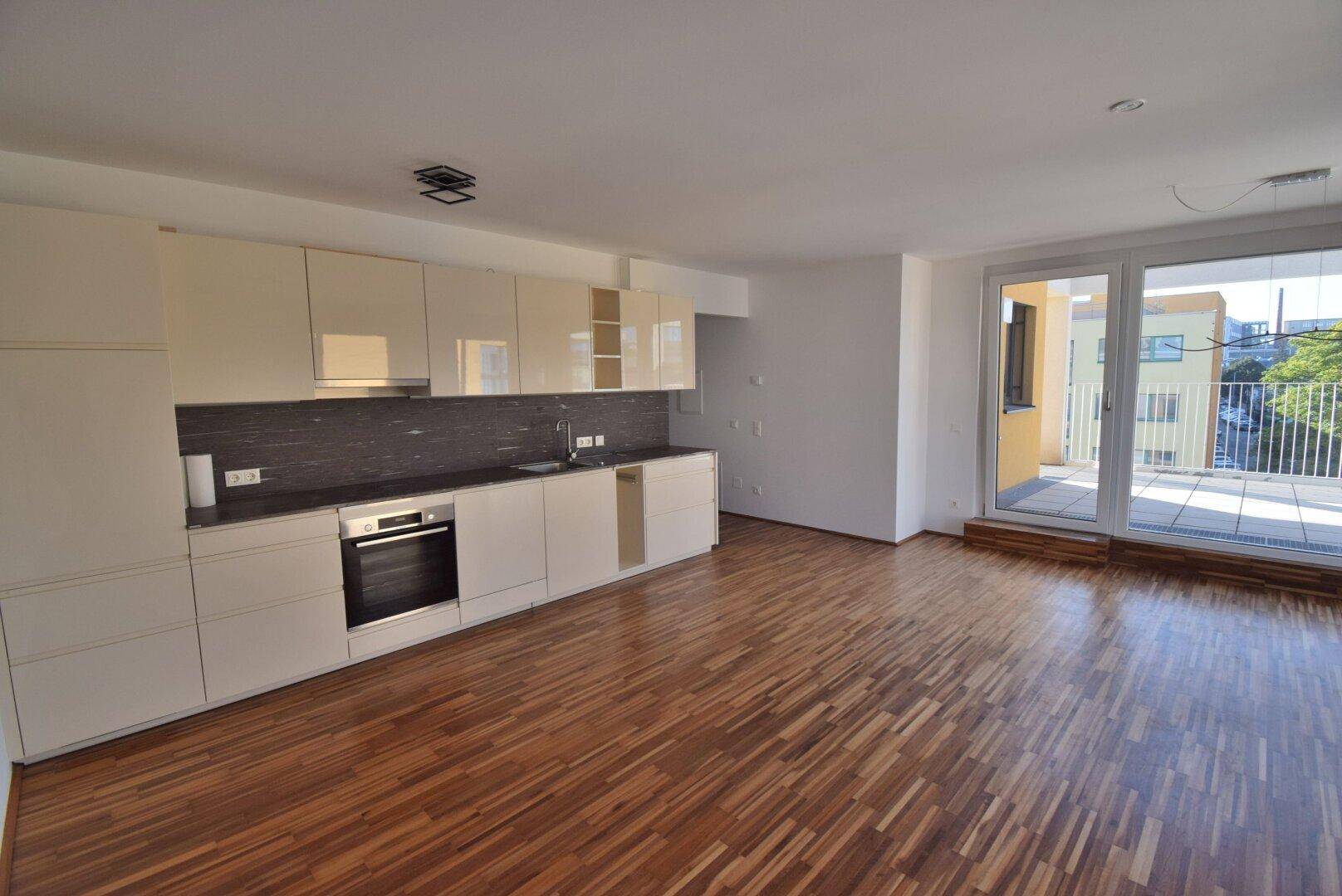 Offener Wohnbereich mit integrierter Küchenzeile, Holzboden und Zugang zum Balkon.
