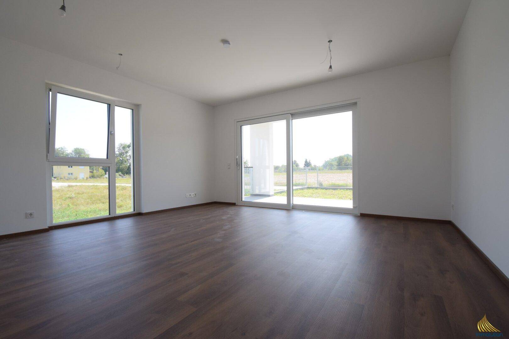 Helles, leeres Wohnzimmer mit großen Fenstern und dunklem Laminatboden, bereit zur Einrichtung.