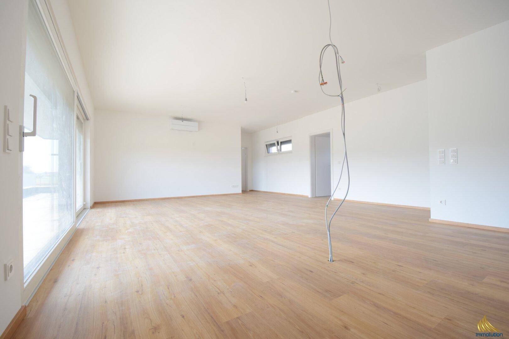 Helles, unmöbliertes Wohnzimmer mit großen Fenstern, Laminatboden und Klimaanlage.