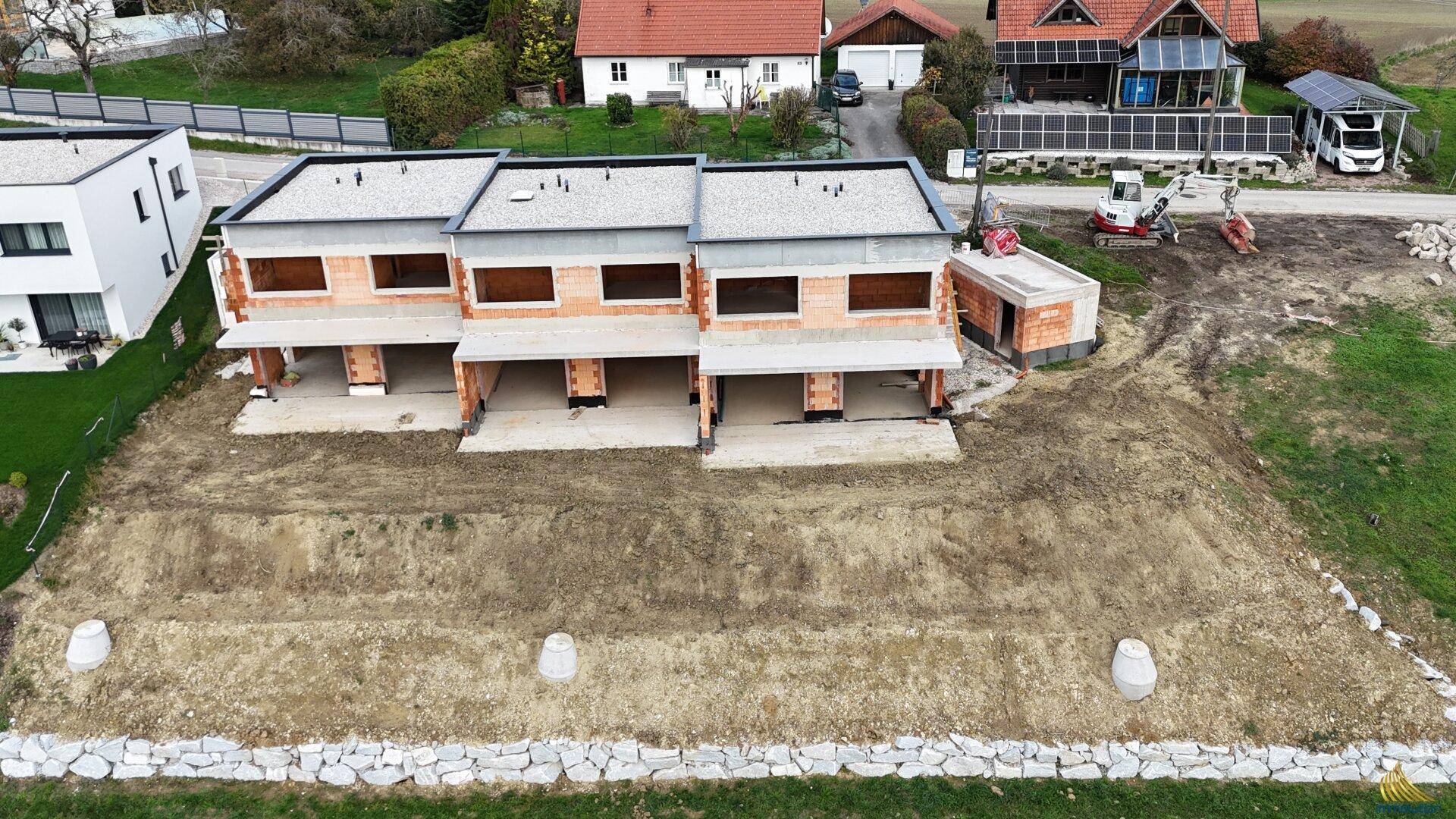 Luftaufnahme einer Baustelle mit drei Reihenhäusern im Rohbauzustand und umliegender Landschaft.