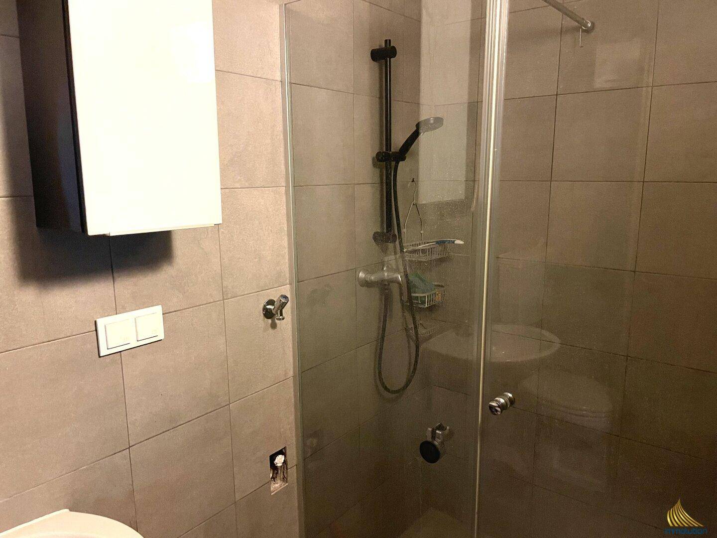 Modernes Badezimmer mit grauen Fliesen, Glasdusche und weißem Hängeschrank.