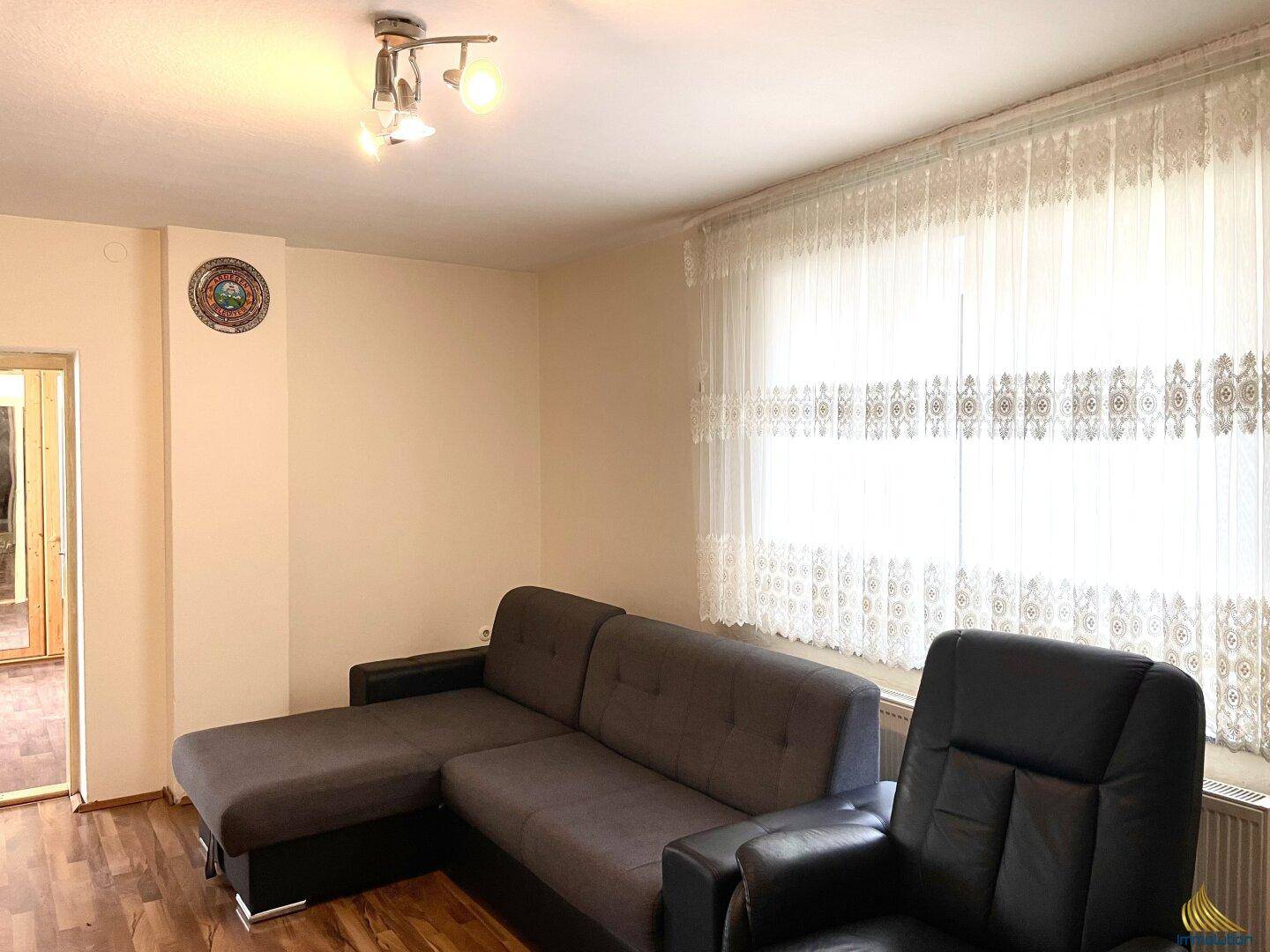 Helles Wohnzimmer mit grauem Ecksofa, Sessel und großen Fenstern mit Vorhängen.