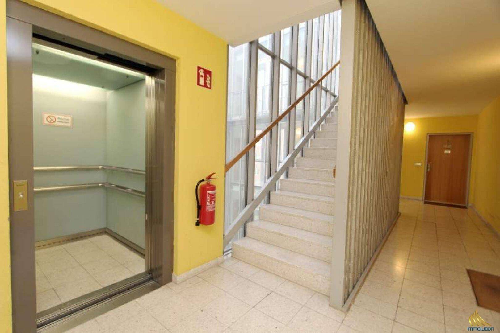 Heller Flur mit Aufzug, Treppenhaus und gelben Wänden, ausgestattet mit einem Feuerlöscher.