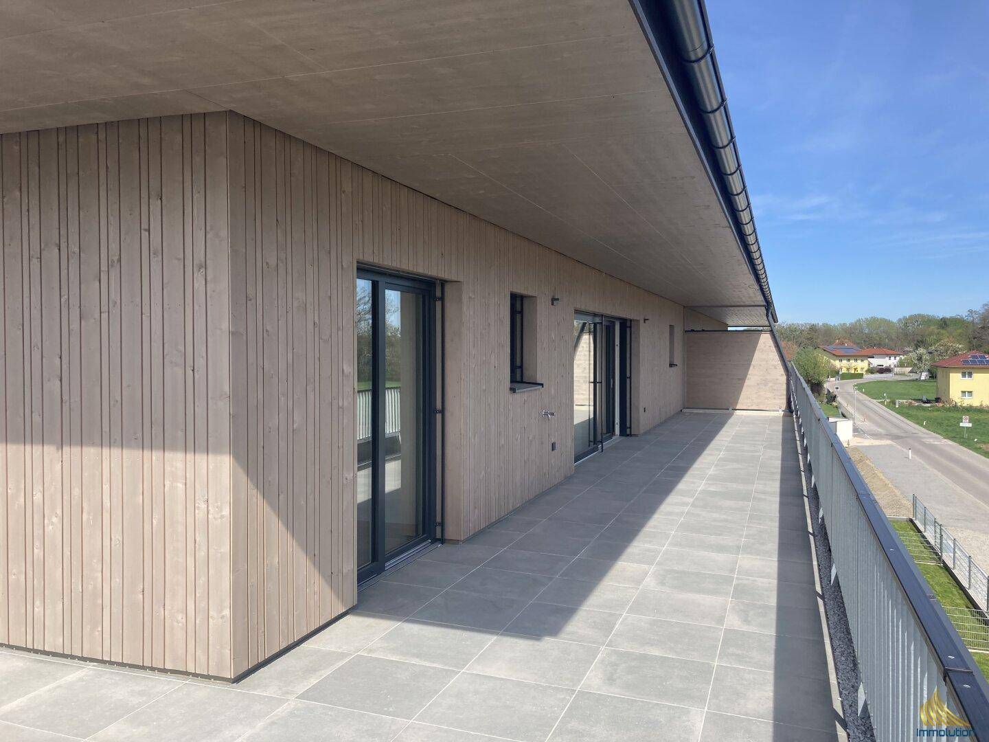 Lange, geflieste Terrasse mit Holzfassade und weitem Blick auf die Umgebung unter blauem Himmel.